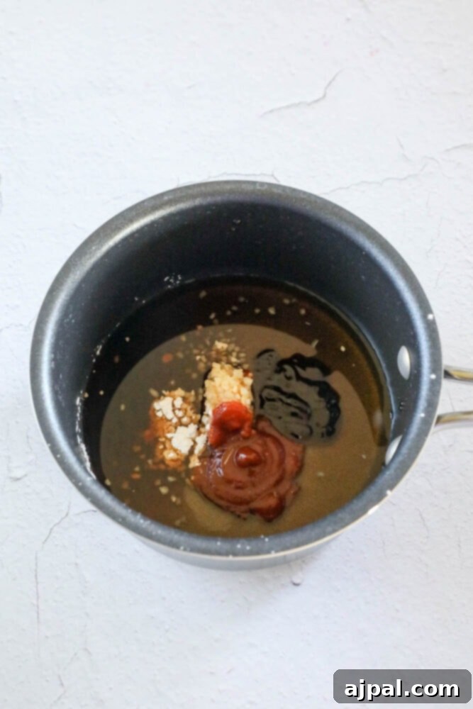 A small saucepan containing sriracha, liquid aminos, sesame seeds, and hoisin sauce, representing the key components for the sweet and spicy glaze.