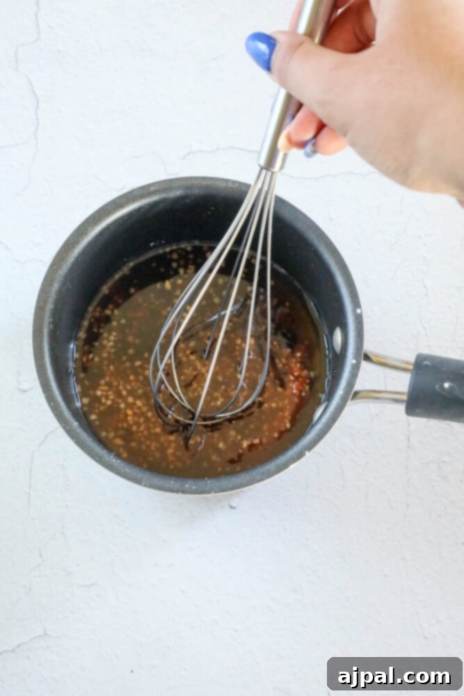 A hand is actively whisking the spicy Asian sauce in a saucepan on the stovetop, demonstrating the preparation of the flavorful glaze.