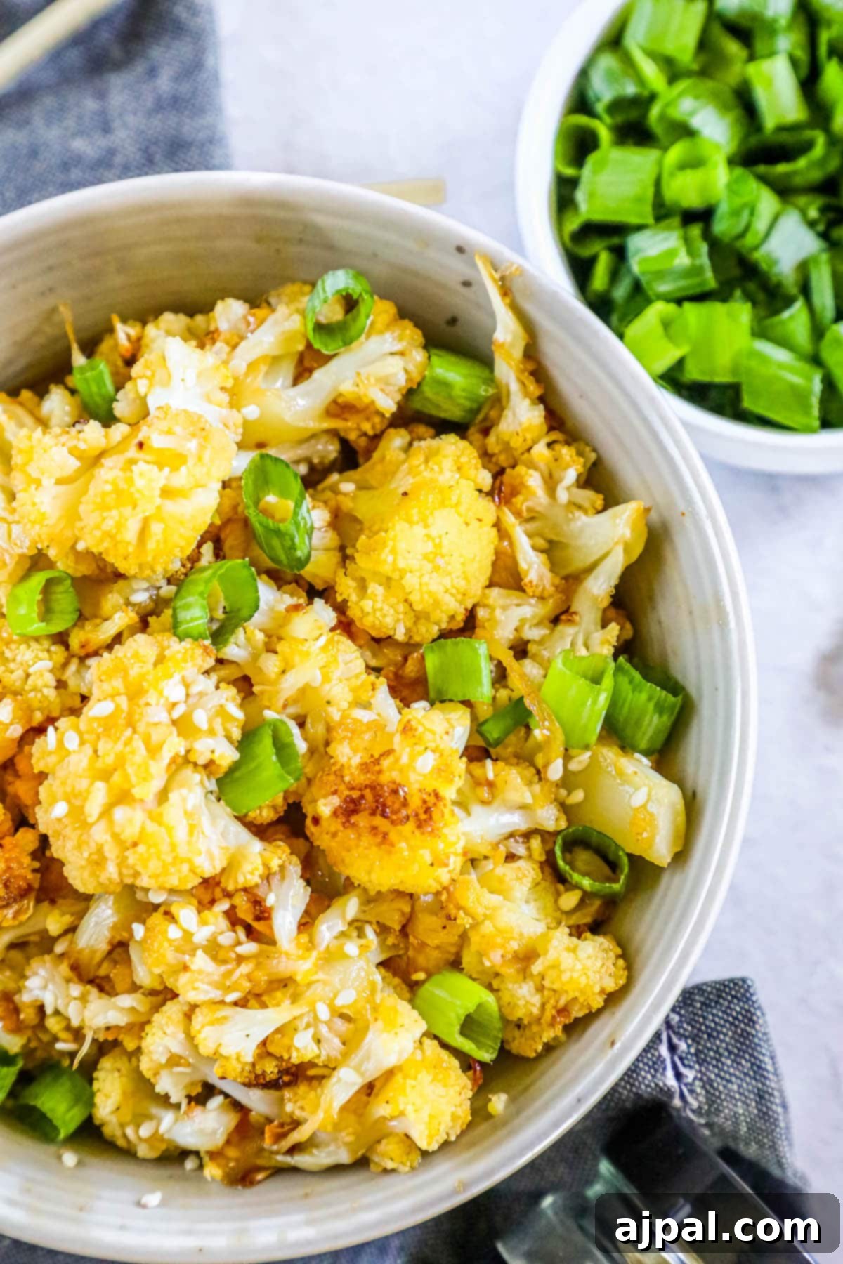 A vibrant bowl of sweet and spicy baked cauliflower, generously topped with sesame seeds and fresh sliced green onions, ready to be served on a table.