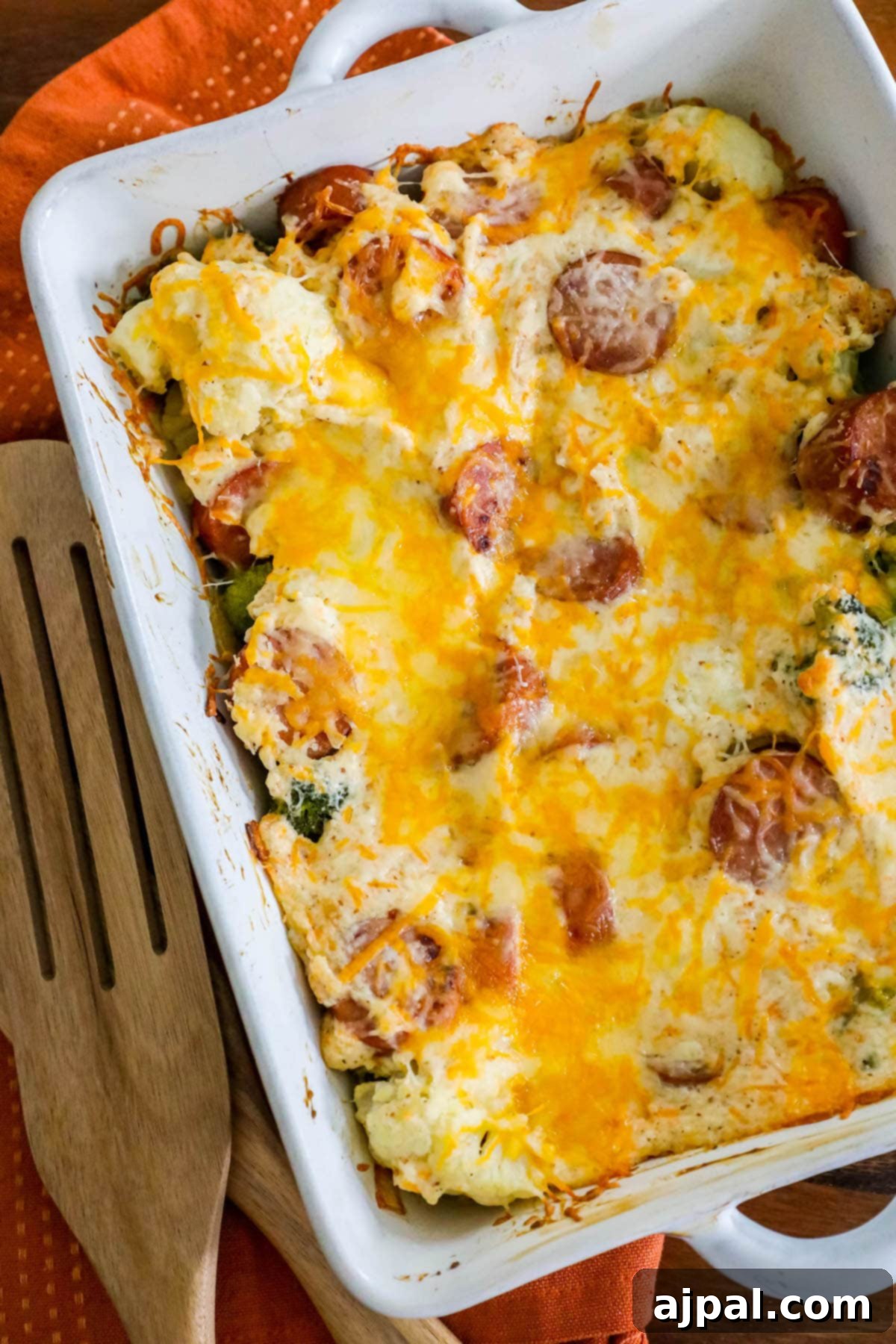 A freshly baked cheesy casserole, featuring sliced sausage, vibrant broccoli florets, and tender cauliflower, elegantly seasoned with herbs, presented in a white baking dish on a rustic wooden table.