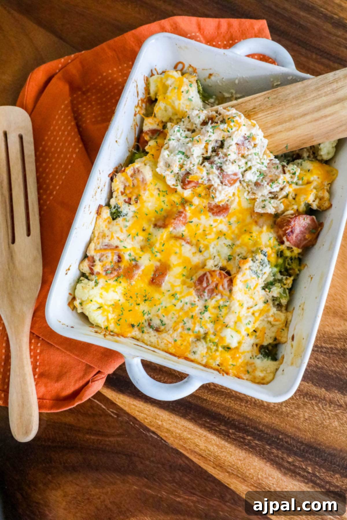 A wooden spoon delicately scooping a generous portion of cheesy casserole, revealing layers of sliced sausage, vibrant broccoli, and tender cauliflower, all seasoned with aromatic herbs, from a pristine white baking dish.