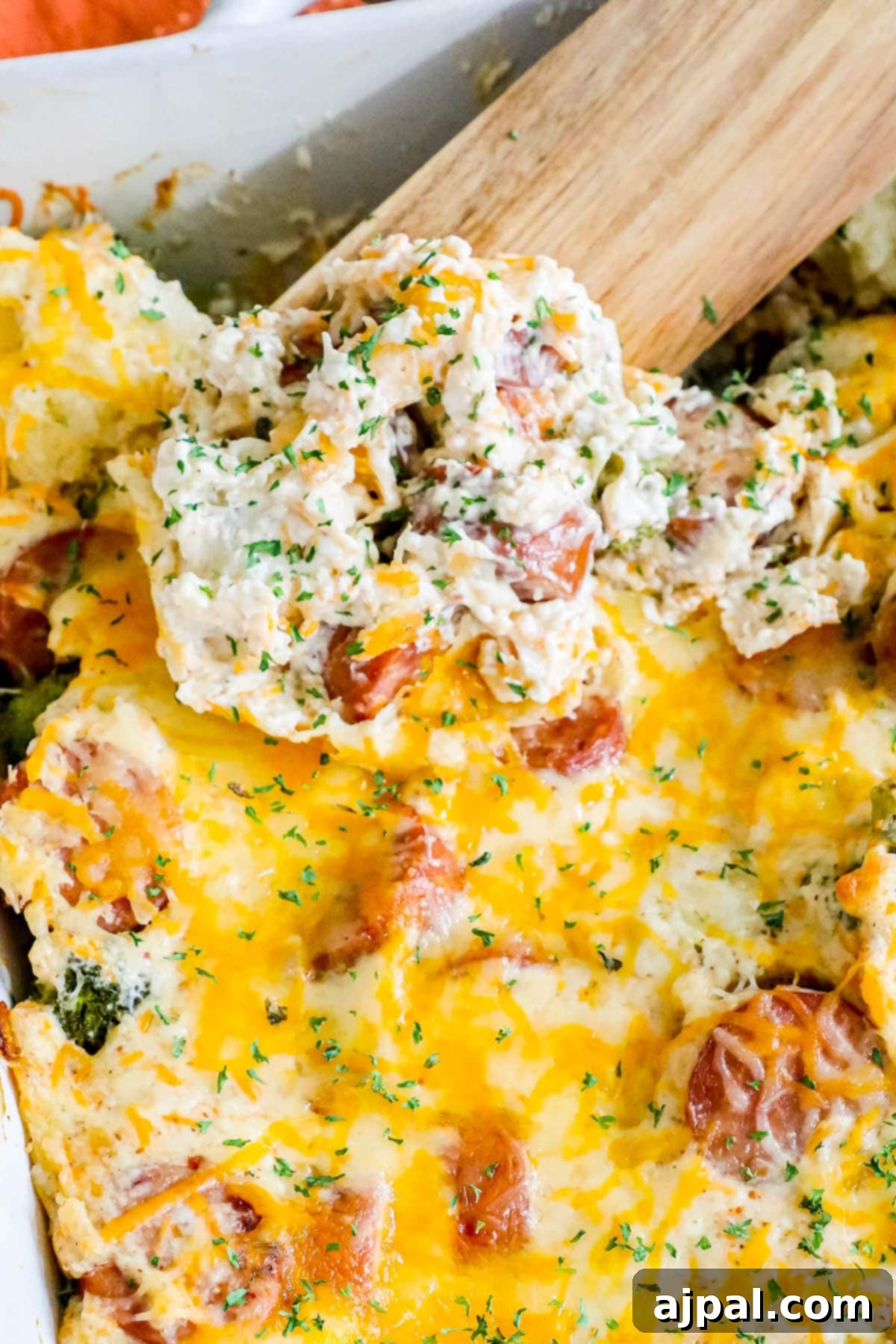 A large wooden spoon dipped into a freshly baked cheesy broccoli cauliflower sausage casserole, showcasing the melty cheese, succulent sausage slices, and perfectly cooked vegetables within the white casserole dish.