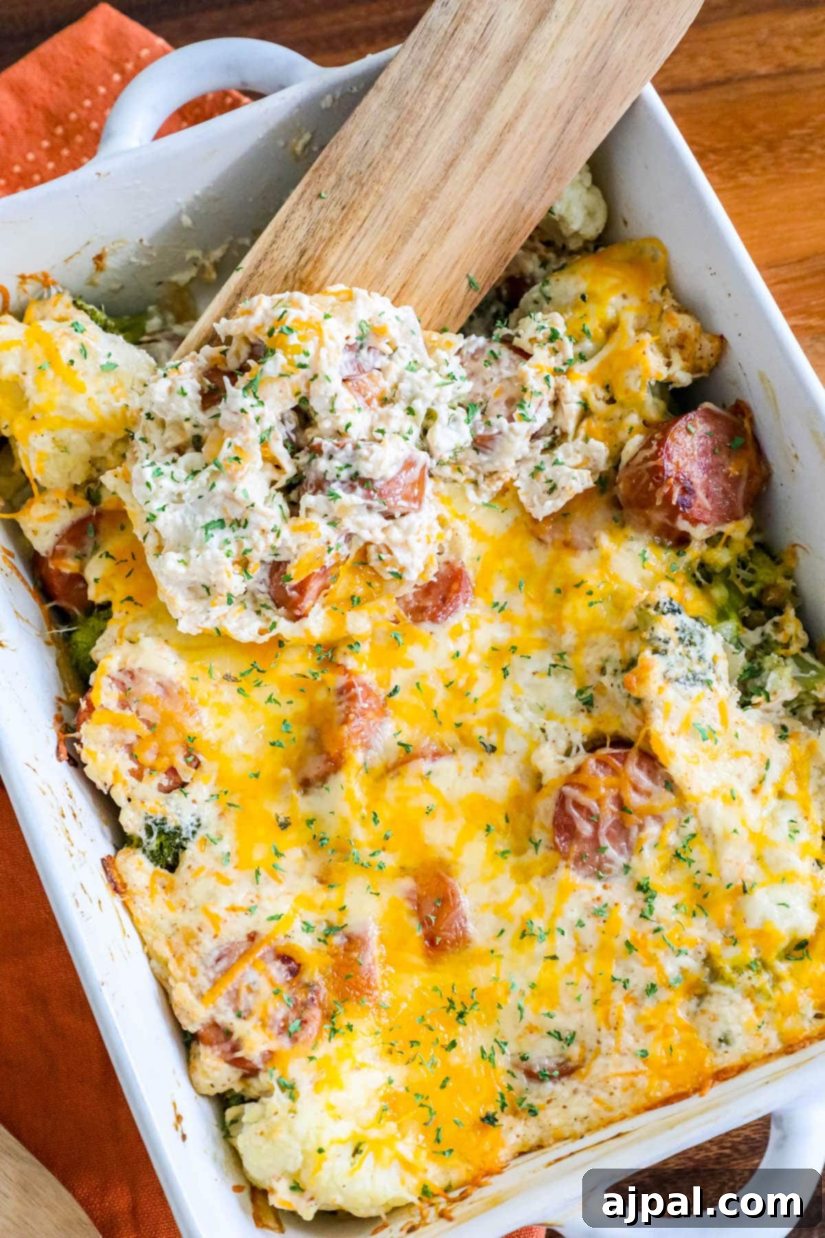 A baked cheesy casserole, brimming with sliced sausage, vibrant broccoli, and tender cauliflower, generously topped with melted cheese and fresh herb seasonings, ready to be served in a white baking dish.