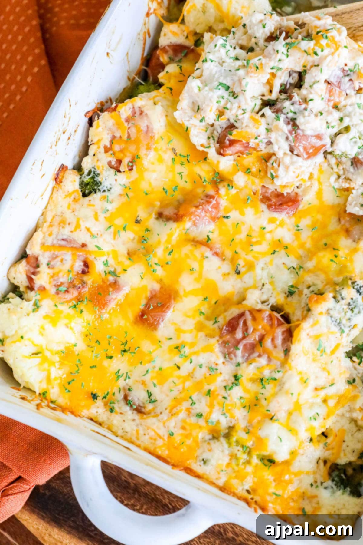 A delectable casserole, featuring perfectly sliced sausage, tender broccoli, and succulent cauliflower, all smothered in rich, melted cheese and garnished with herbs, presented in a pristine white baking dish on a wooden table.