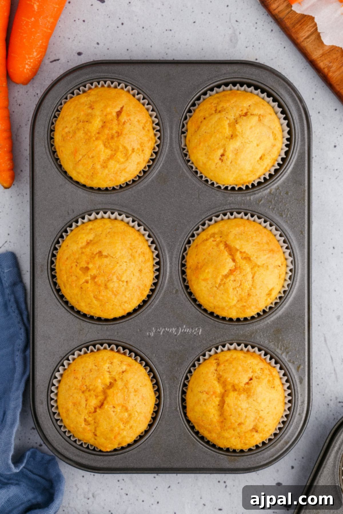 Effortless Carrot Muffins 2 6 freshly baked carrot muffins in a muffin tin, surrounded by vibrant raw carrots, ready to be enjoyed.