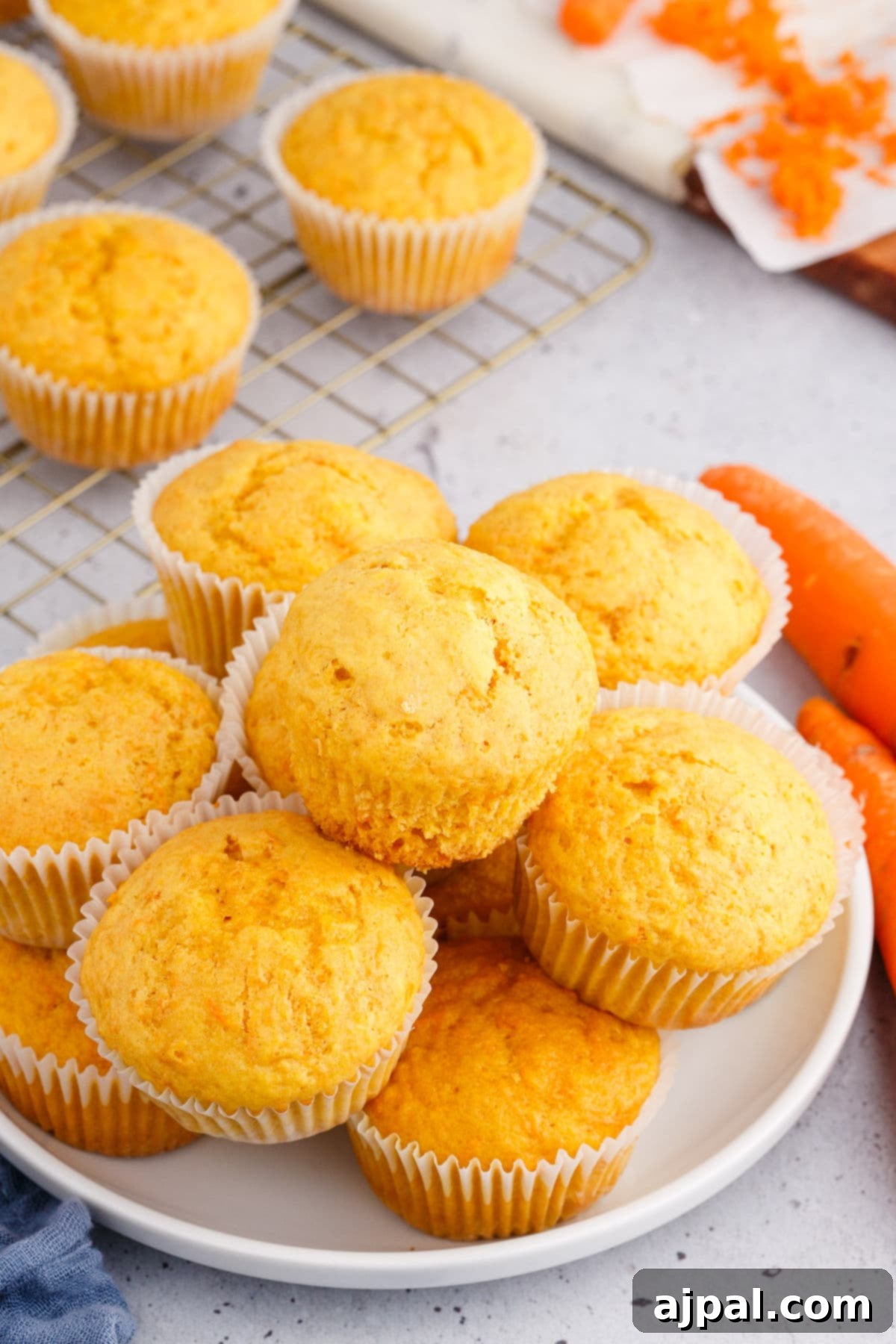 Effortless Carrot Muffins 14 Carrot muffins with white cupcake liners, elegantly stacked on a pristine white plate, accompanied by fresh carrots.