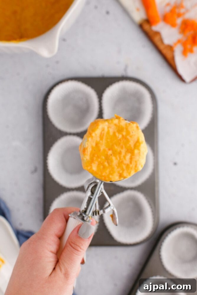 Effortless Carrot Muffins 10 A hand expertly holds a metal cupcake scooper, brimming with carrot muffin batter, poised above a muffin tin meticulously lined with cupcake wrappers.
