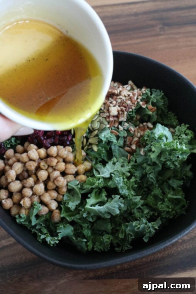 Jeweled Chickpea Pomegranate Salad 7 a hand pouring dressing over a salad of chickpeas, kale, dried cranberries, pomegranate arils, pepitas, and sunflower seeds
