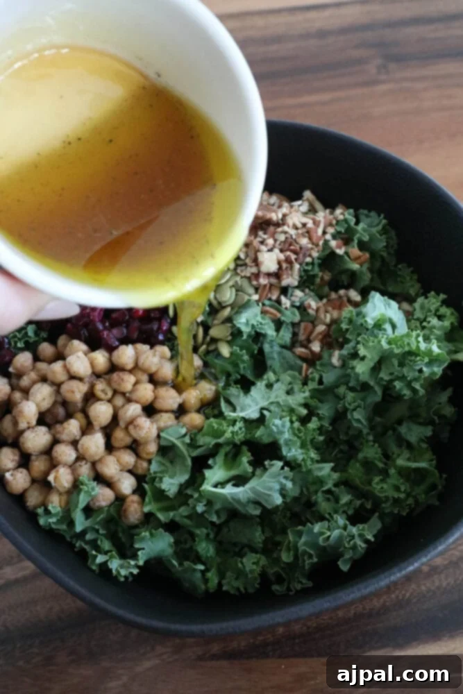 Mediterranean Chickpea Pomegranate Salad 7 a hand pouring dressing over a salad of chickpeas, kale, dried cranberries, pomegranate arils, pepitas, and sunflower seeds