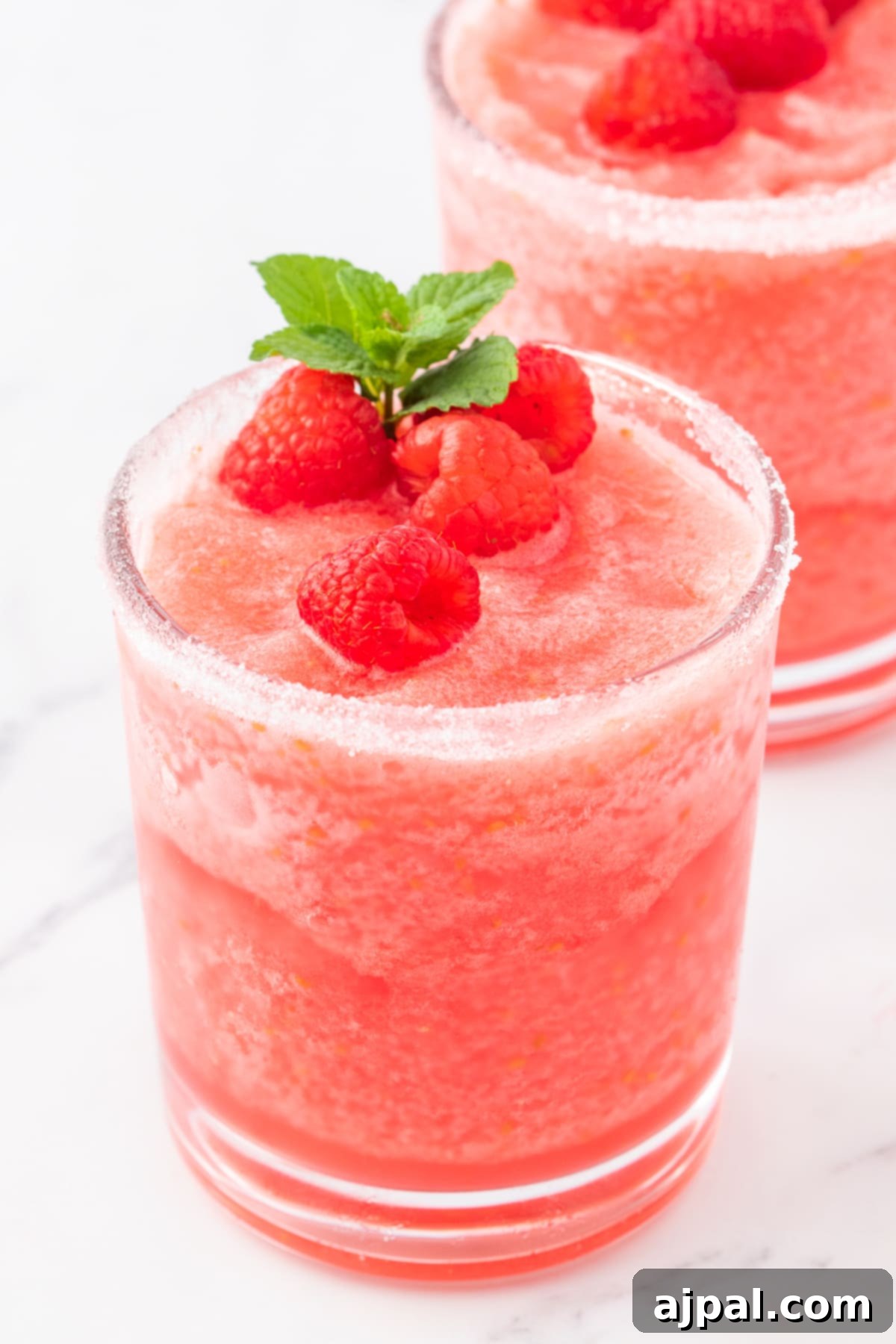two glasses of frozen pink lemonade in a sugar rimmed glass with raspberries and a sprig of mint on top