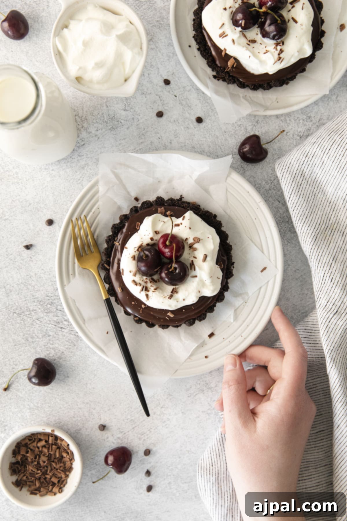 Dark Forest Cherry Delights 3 A close-up shot of a hand reaching for a plate, where two Black Forest tarts are displayed. Each tart features a chocolate ganache filling in an Oreo crust, lavishly topped with whipped cream and gleaming fresh cherries, inviting indulgence.