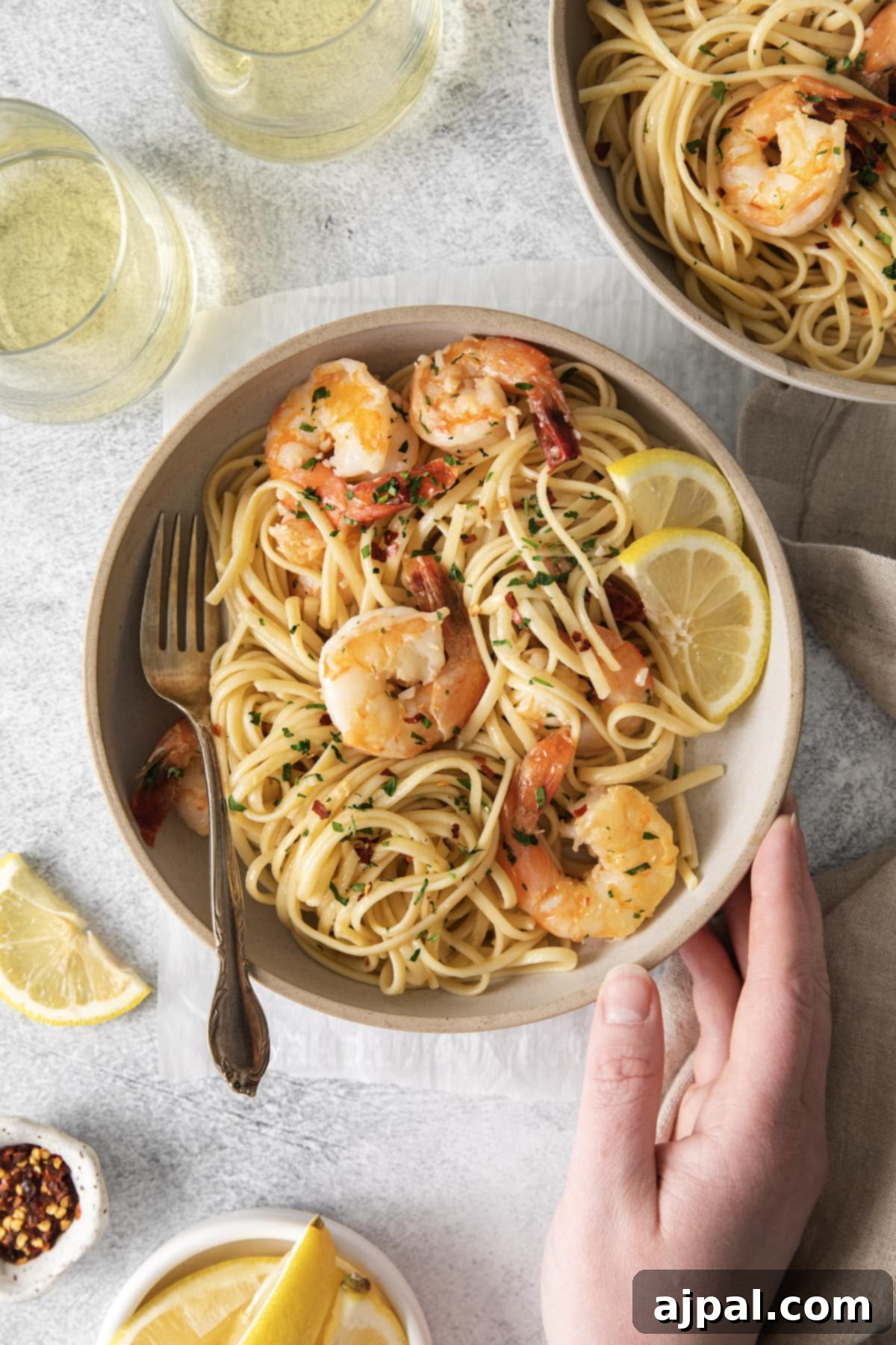 Effortless Shrimp Scampi 3 a hand touching a bowl with shrimp pasta with linguine, red pepper flakes, herbs, and lemon slices