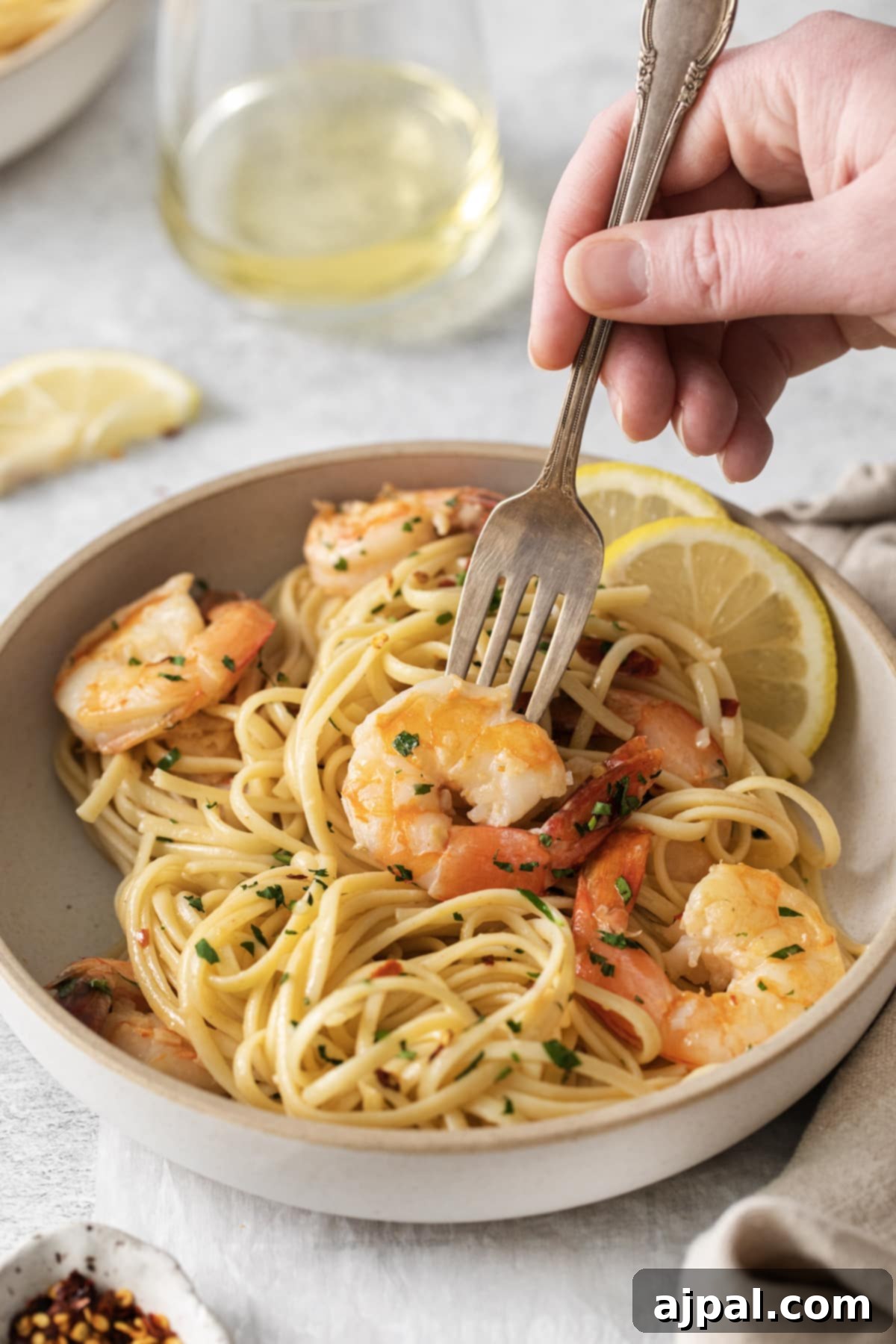 Effortless Shrimp Scampi 10 a hand holding a fork twirling shrimp pasta with linguine, red pepper flakes, herbs, and lemon slices in a bowl
