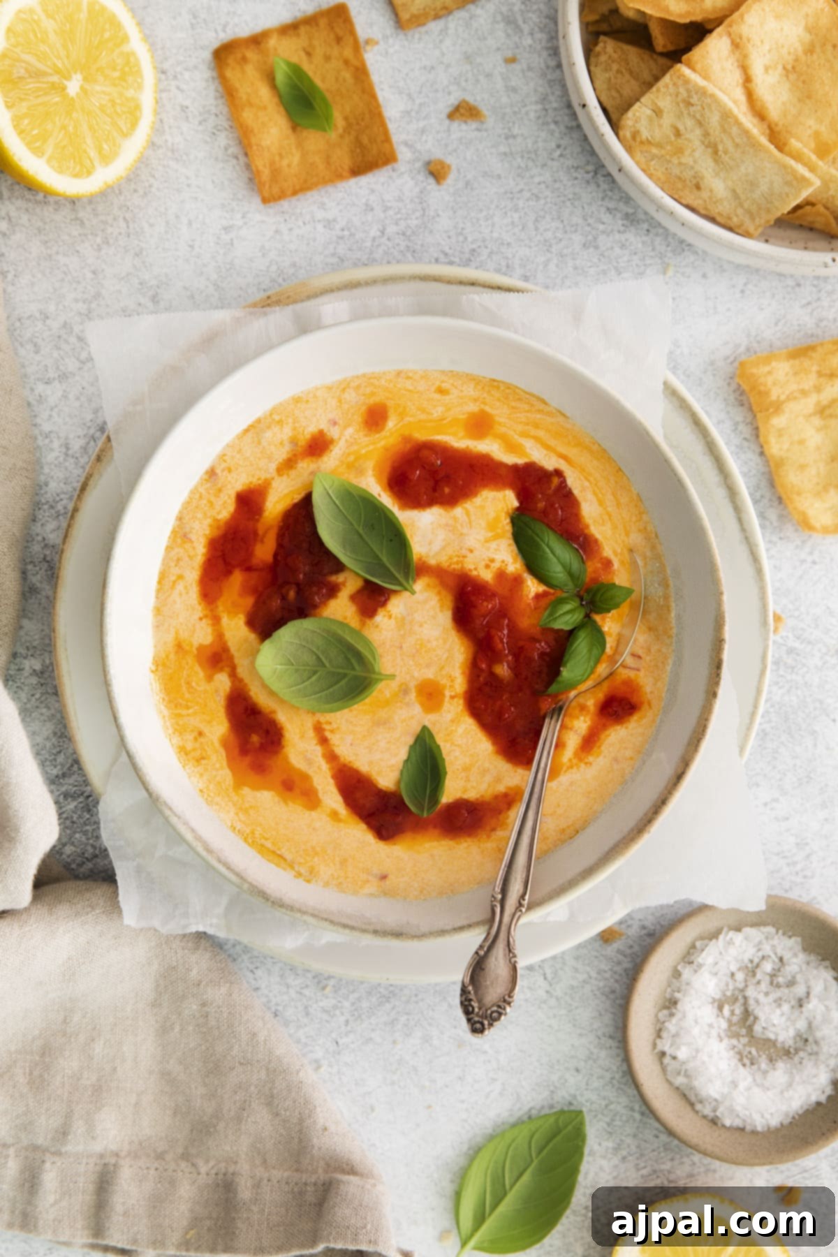 Creamy whipped feta in a bowl, garnished with fresh basil sprigs and a spoon in the middle. Swirls of harissa paste add a vibrant touch.