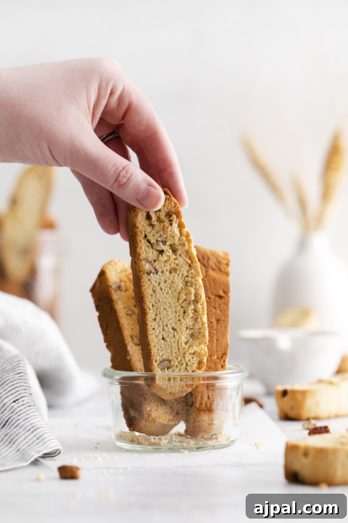 Golden Maple Pecan Biscotti 2 A hand reaching for a freshly baked maple pecan biscotti with whole pecans in a glass jar on a rustic wooden table.