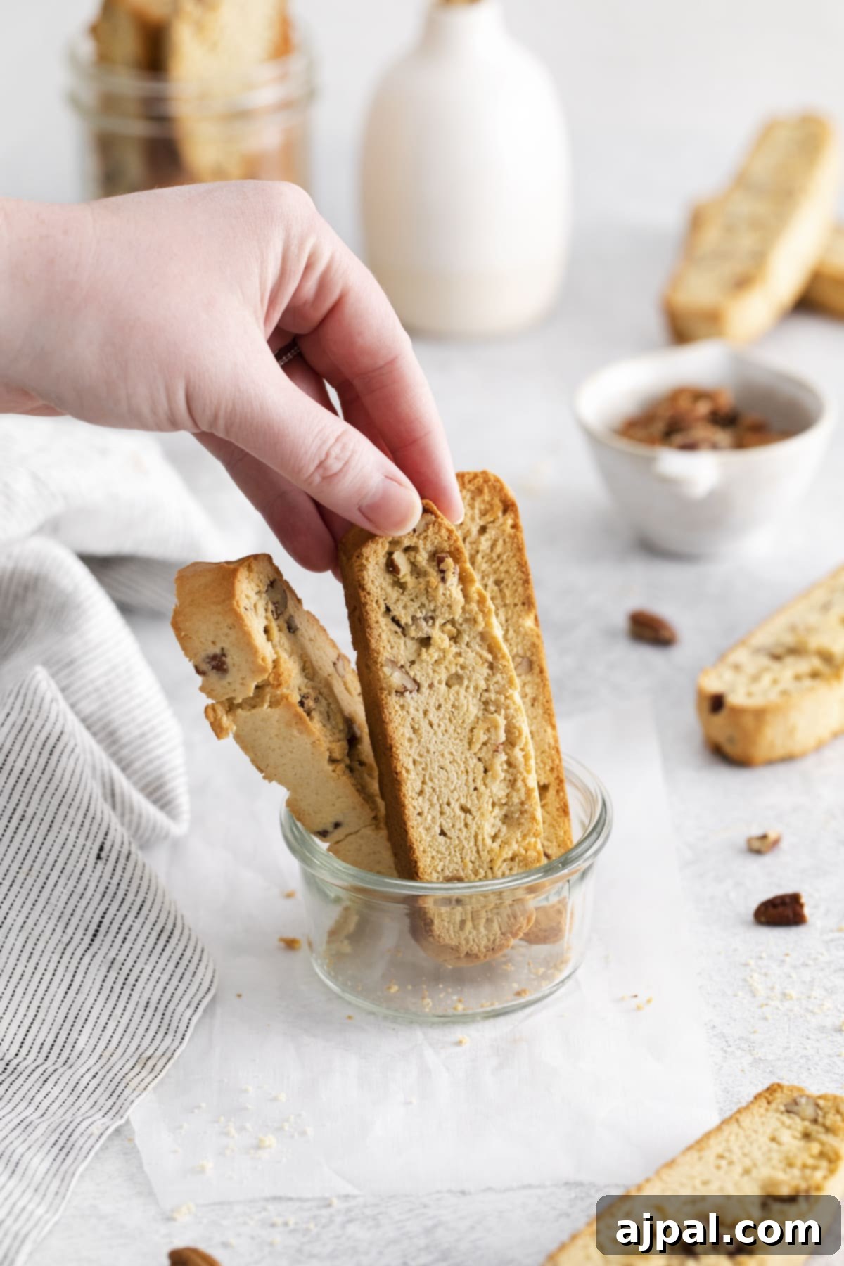 Golden Maple Pecan Biscotti 11 A person holding a piece of maple pecan biscotti, about to dip it into a cup of coffee or tea. A jar of biscotti is visible in the background.