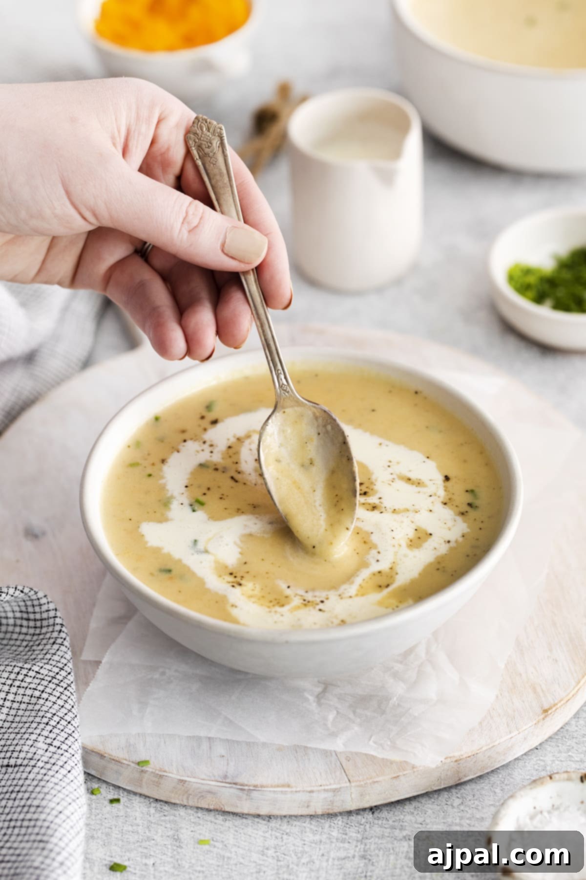 A hand gently dips a spoon into a creamy bowl of apple cheddar soup, highlighting the rich texture and inviting garnish of cream and cracked pepper on top.