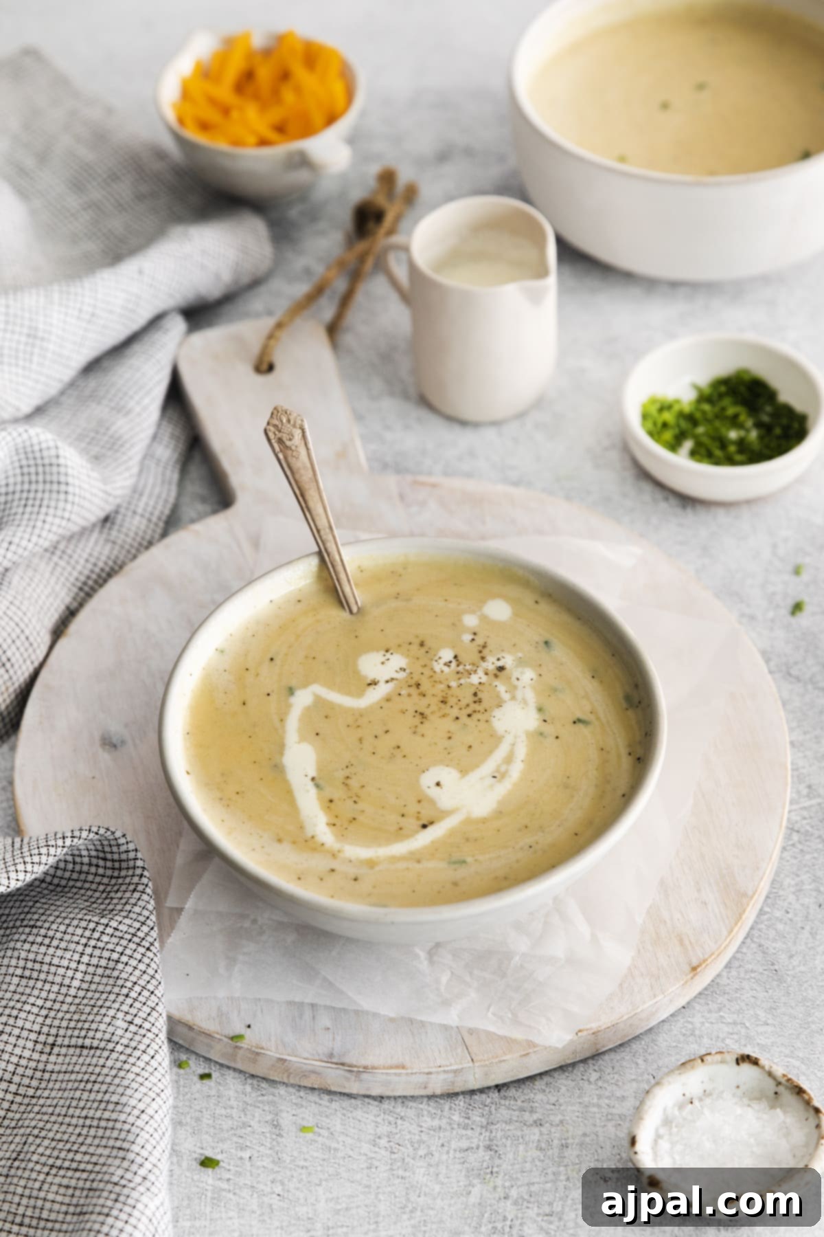 A spoon is nestled in a rich bowl of apple cheddar soup, garnished with a swirl of cream and a crack of pepper, placed on a simple wooden cutting board.