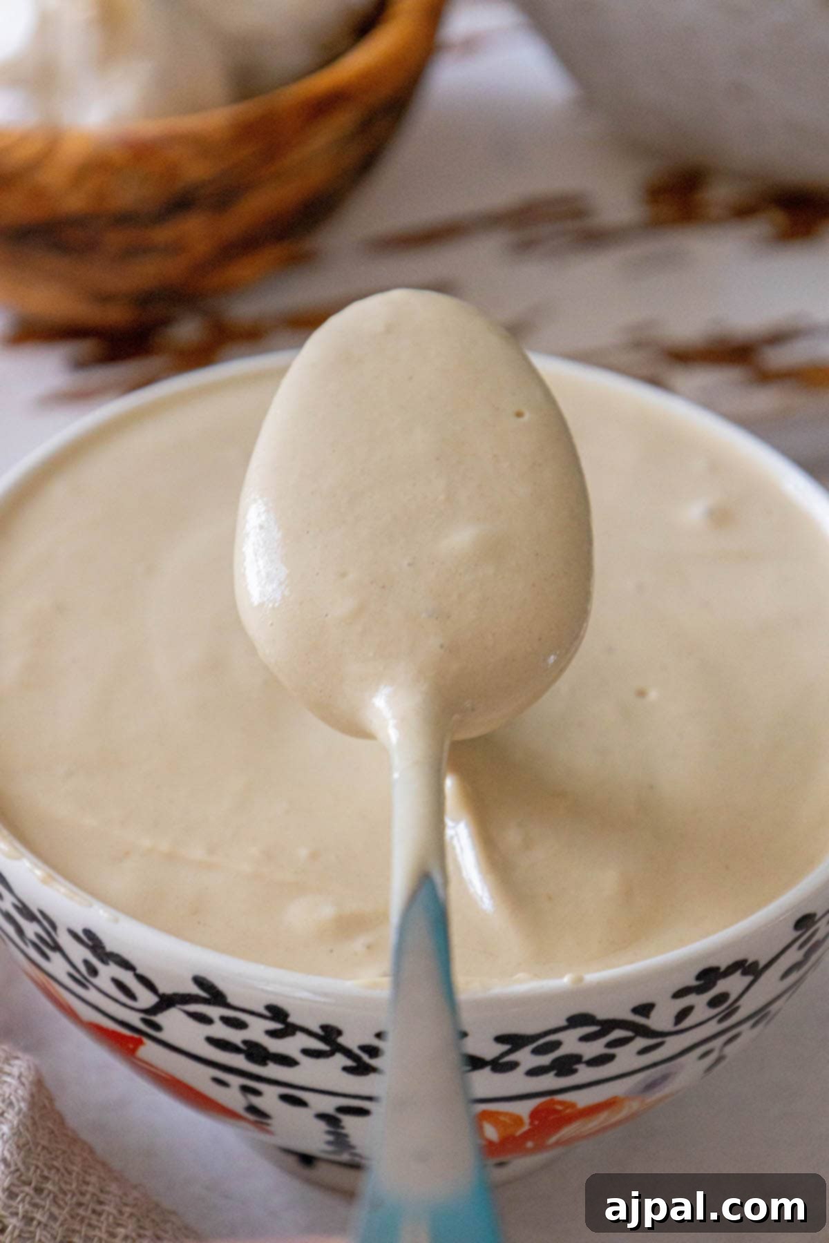 A spoon delicately lifting creamy tahini sauce over a bowl on a rustic wooden table.