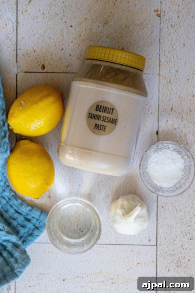 Fresh ingredients laid out: tahini paste, vibrant lemons, a bowl of cold water, a pinch of salt, and fragrant garlic cloves, ready for mixing.