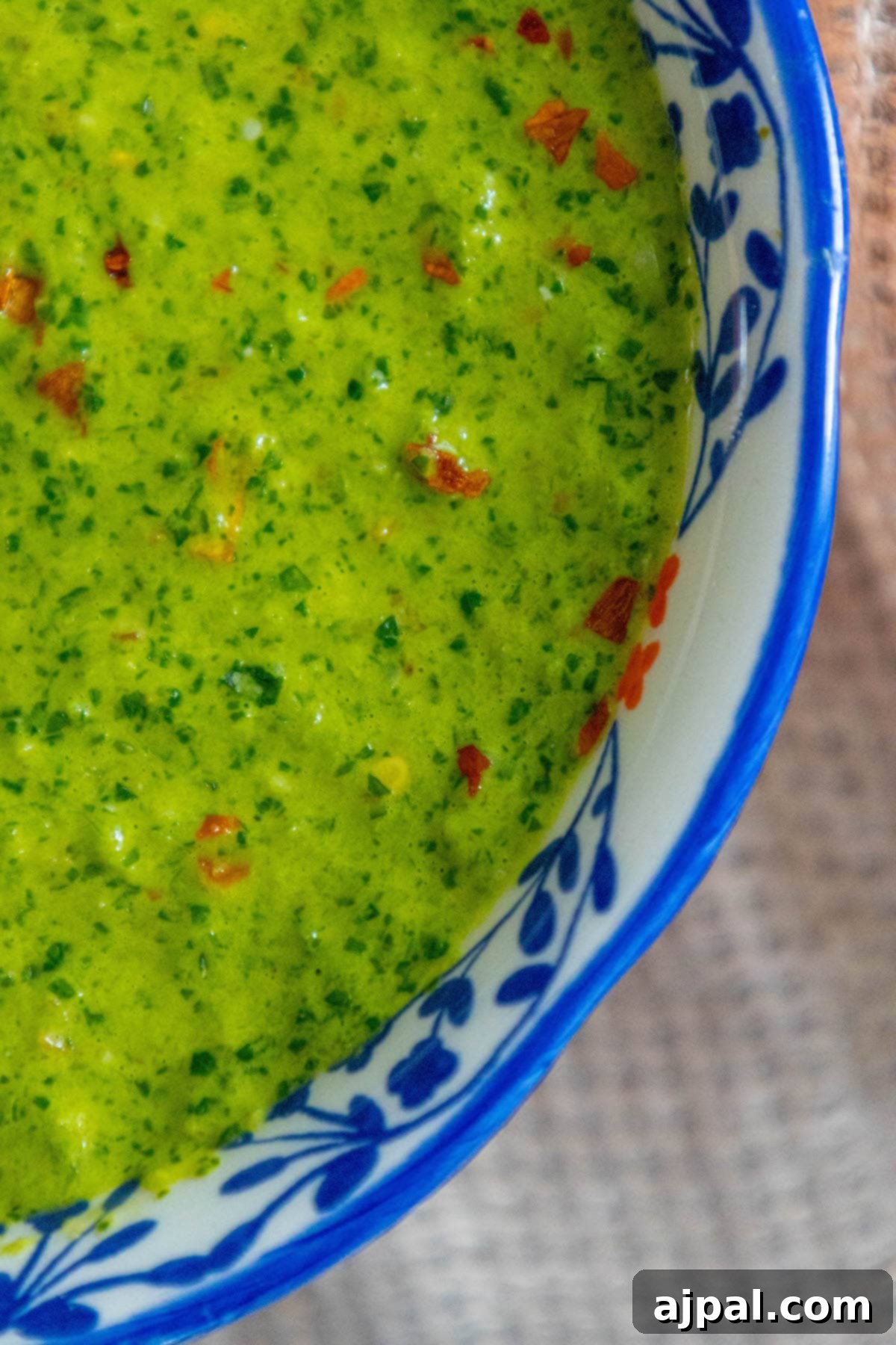 Skhug Zhoug Sauce Recipe 2 Bright green zhoug sauce with delicate flecks of cilantro and vibrant red pepper flakes, artfully presented in a decorative blue mug, inviting you to spoon it over your next meal.