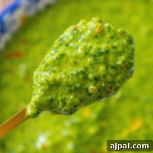 Bright green sauce with flecks of fresh cilantro and vibrant red pepper flakes, beautifully balanced on a spoon above a bowl of zhoug sauce, showcasing its appealing texture and rich color.