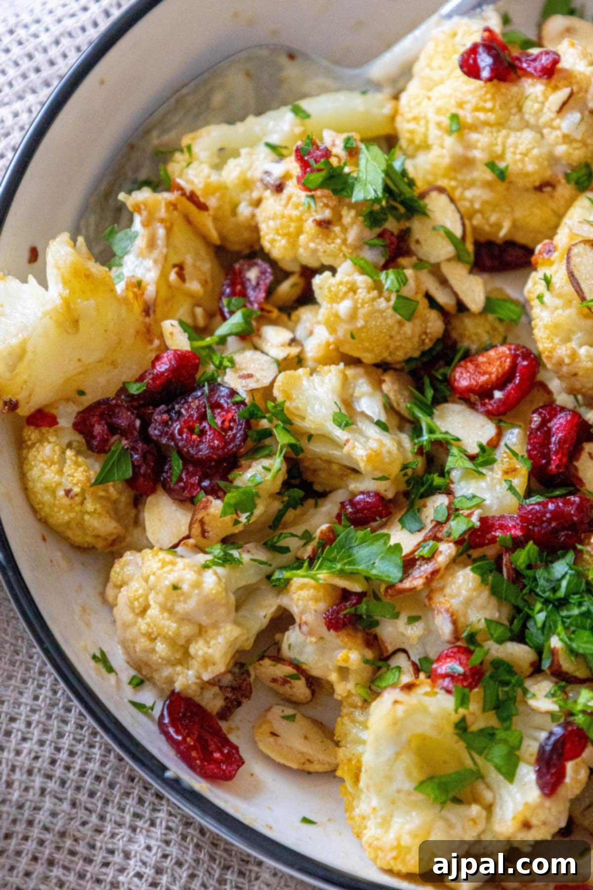 A close-up of roasted cauliflower tossed in tahini sauce with cranberries, almonds, and chopped parsley in a white dish, showcasing its appealing texture and garnish.