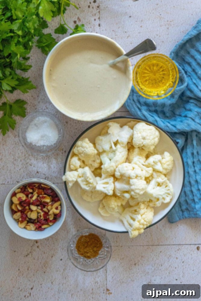 A collection of fresh ingredients for tahini roasted cauliflower: tahini sauce, olive oil, almonds, cranberries, cauliflower, salt, and herbs neatly arranged on a wooden table.