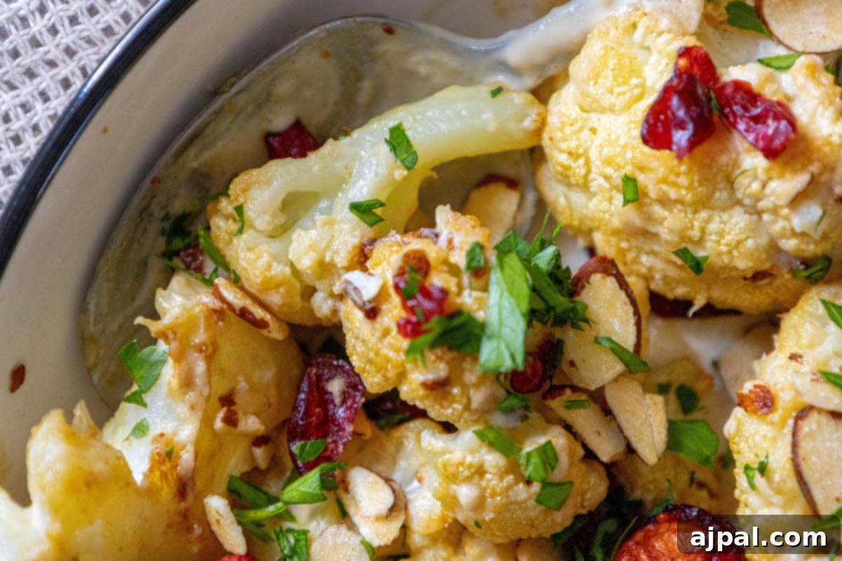 A wide shot of roasted cauliflower tossed in tahini sauce with cranberries, almonds, and chopped parsley in a white dish, presented beautifully.