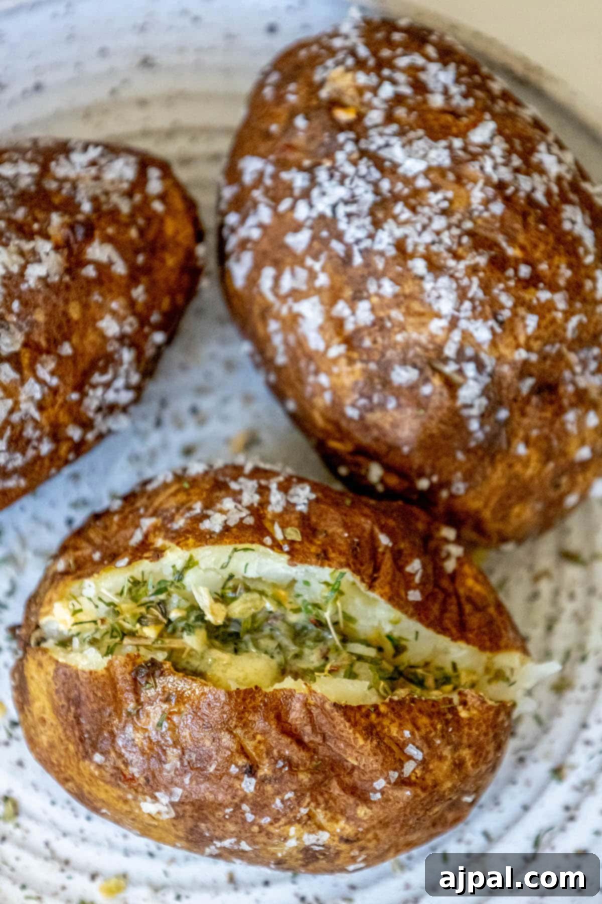 baked potato covered in flaky sea salt cut in half with butter in the middle and herb seasoning inside on a speckled plate with two other salt baked potatoes on the plate next to it
