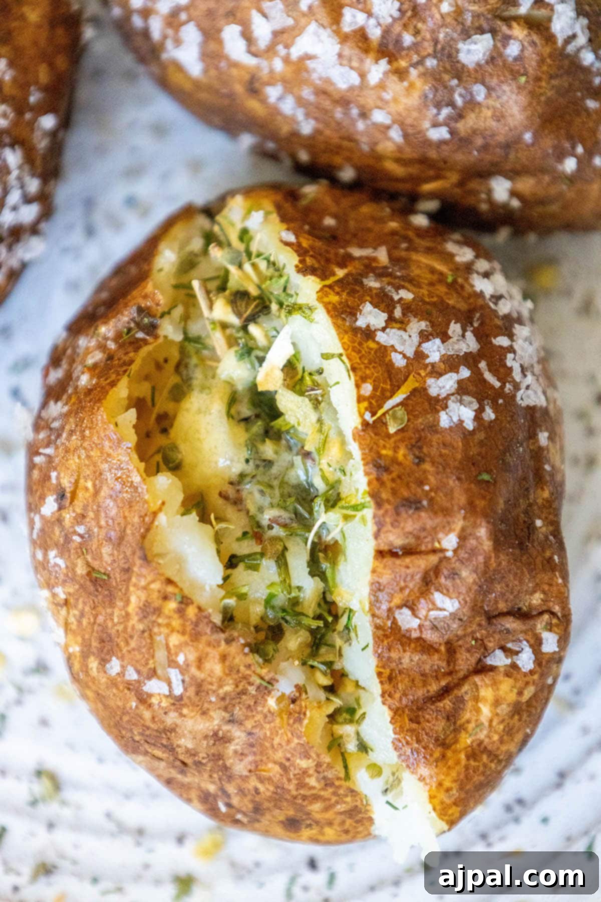 baked potato covered in flaky sea salt cut in half with butter in the middle and herb seasoning inside on a speckled plate with two other salt baked potatoes on the plate next to it