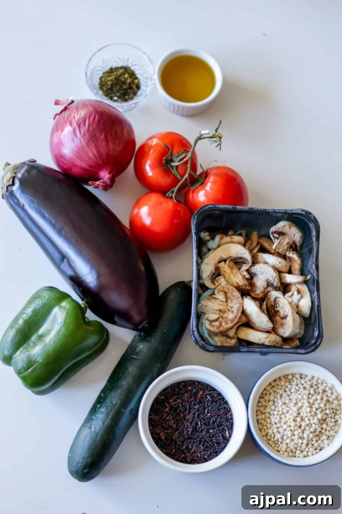 Assorted fresh vegetables including eggplant, red onion, tomatoes, green pepper, cucumber, and mushrooms elegantly arranged in bowls alongside olive oil, herbs, black rice, and couscous on a white surface. Perfect for creating a delightful vegan feast.
