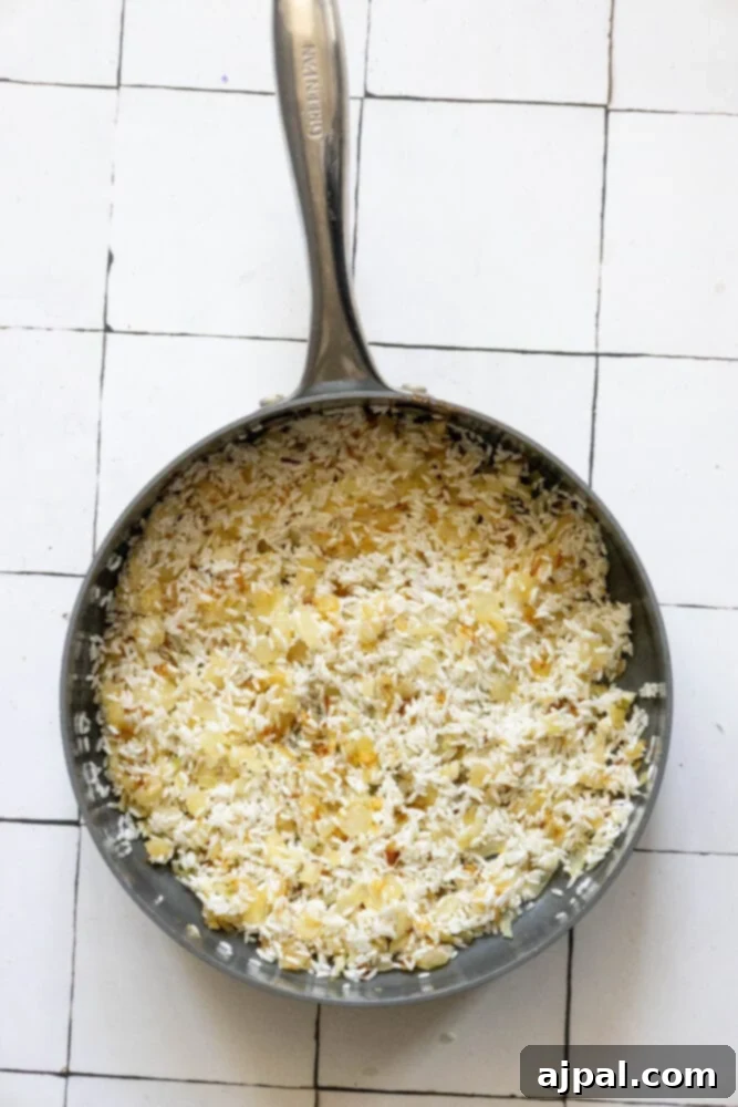 Raw long-grain rice, alongside softened onions and garlic, lightly toasting in a pan, beginning to develop a golden hue.