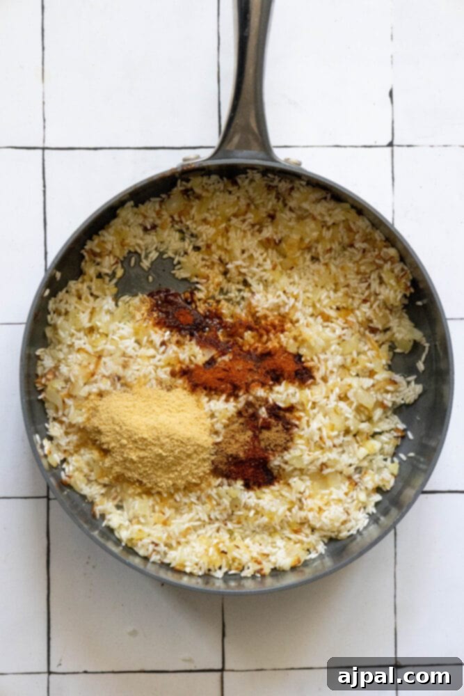 A mix of toasted rice, sautéed onions, and garlic in a pan, with a generous amount of colorful spices freshly added on top, ready to be stirred in.