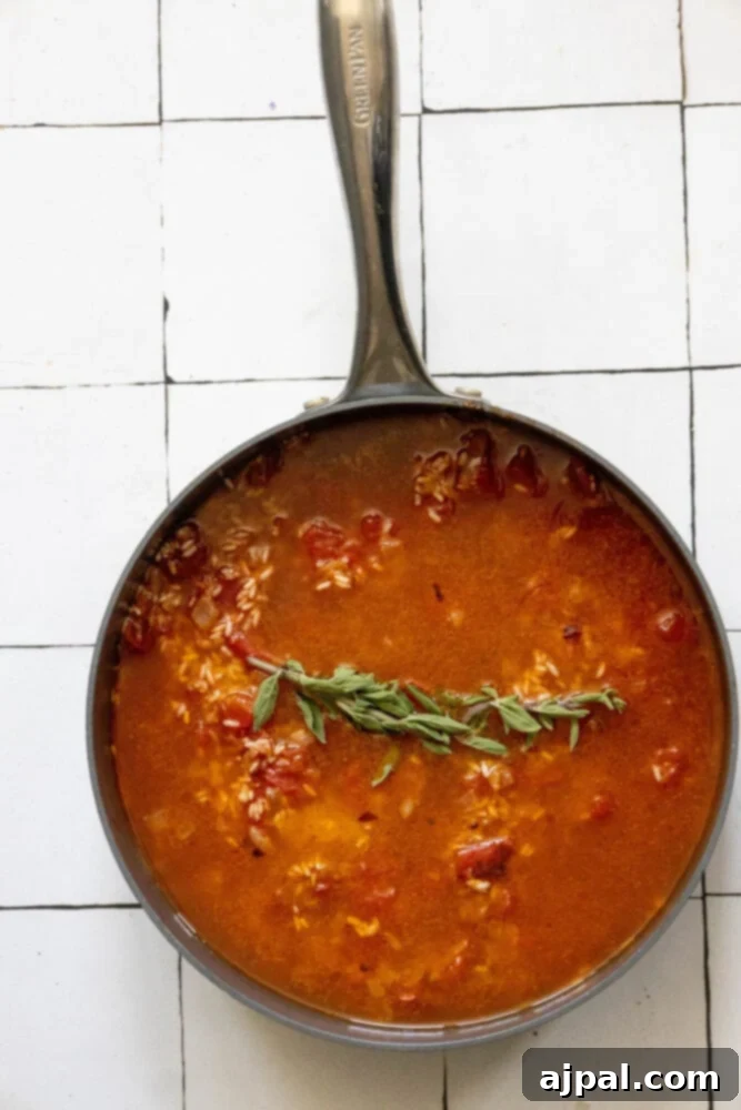 A large skillet filled with long-grain rice, diced tomatoes, herbs, and simmering broth, all combined and ready to cook into Mexican rice.