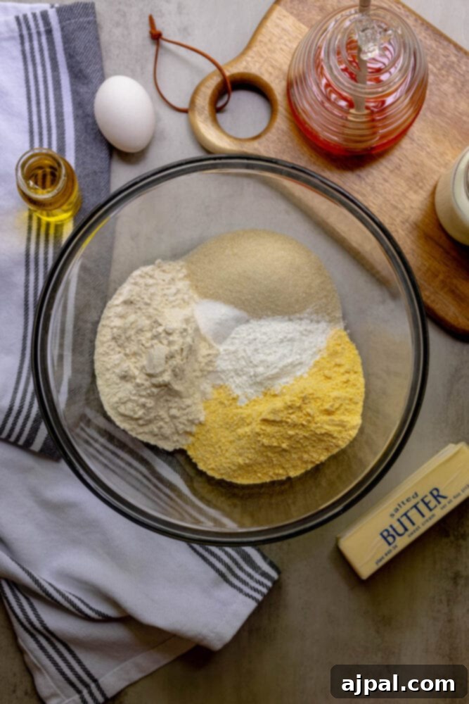 The dry ingredients for cornbread, a blend of flour, cornmeal, sugar, salt, and baking powder, are neatly combined in a clear glass bowl.