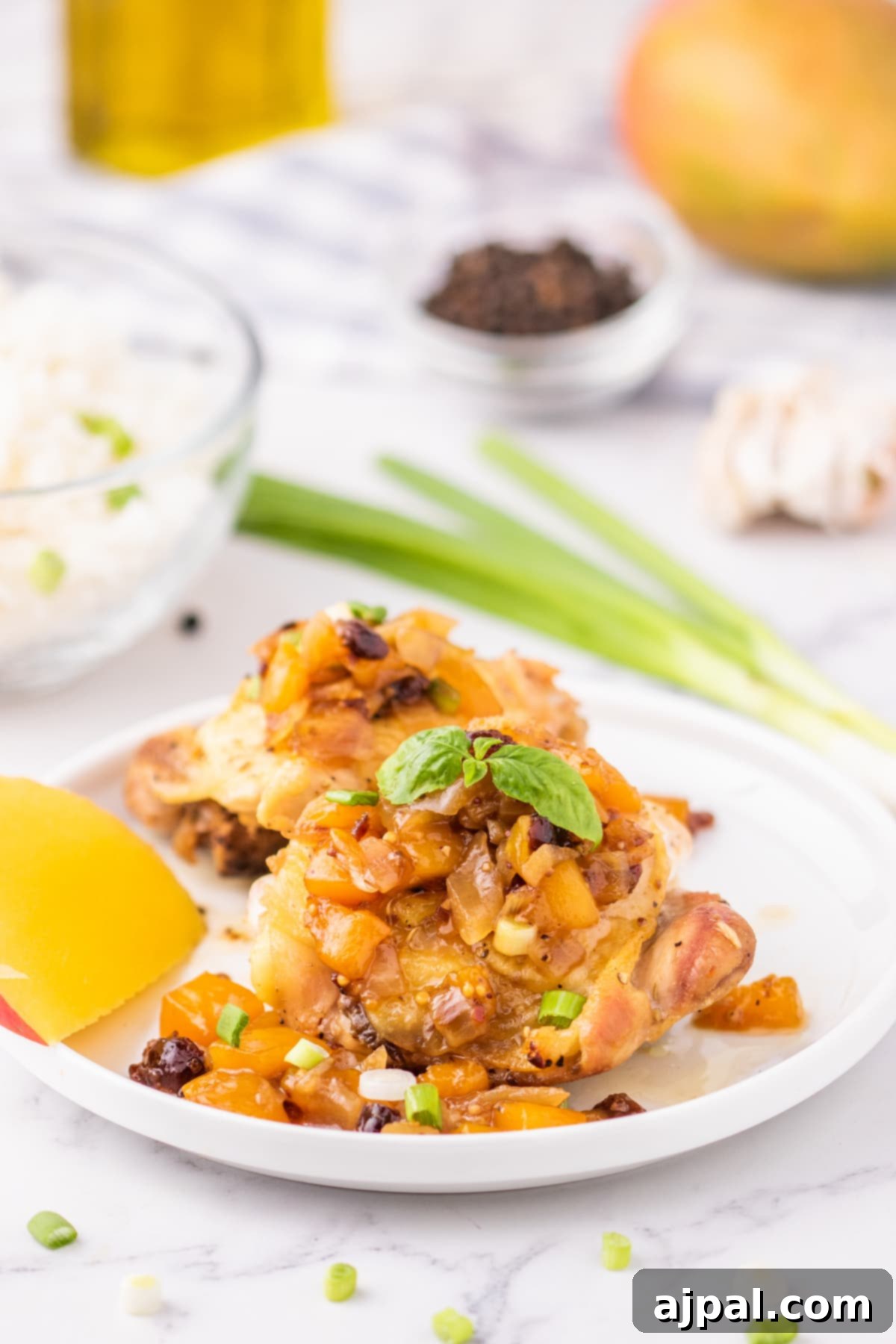 Golden baked chicken thigh on a white plate, generously covered in glistening mango chutney and garnished with fresh, finely sliced green onions.