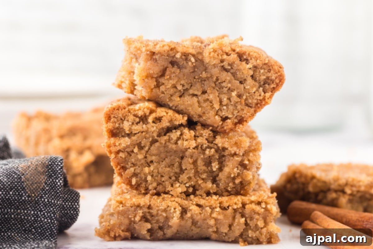 A stack of golden Snickerdoodle Blondies, chewy and warm, adorned with cinnamon sticks on a wooden surface.