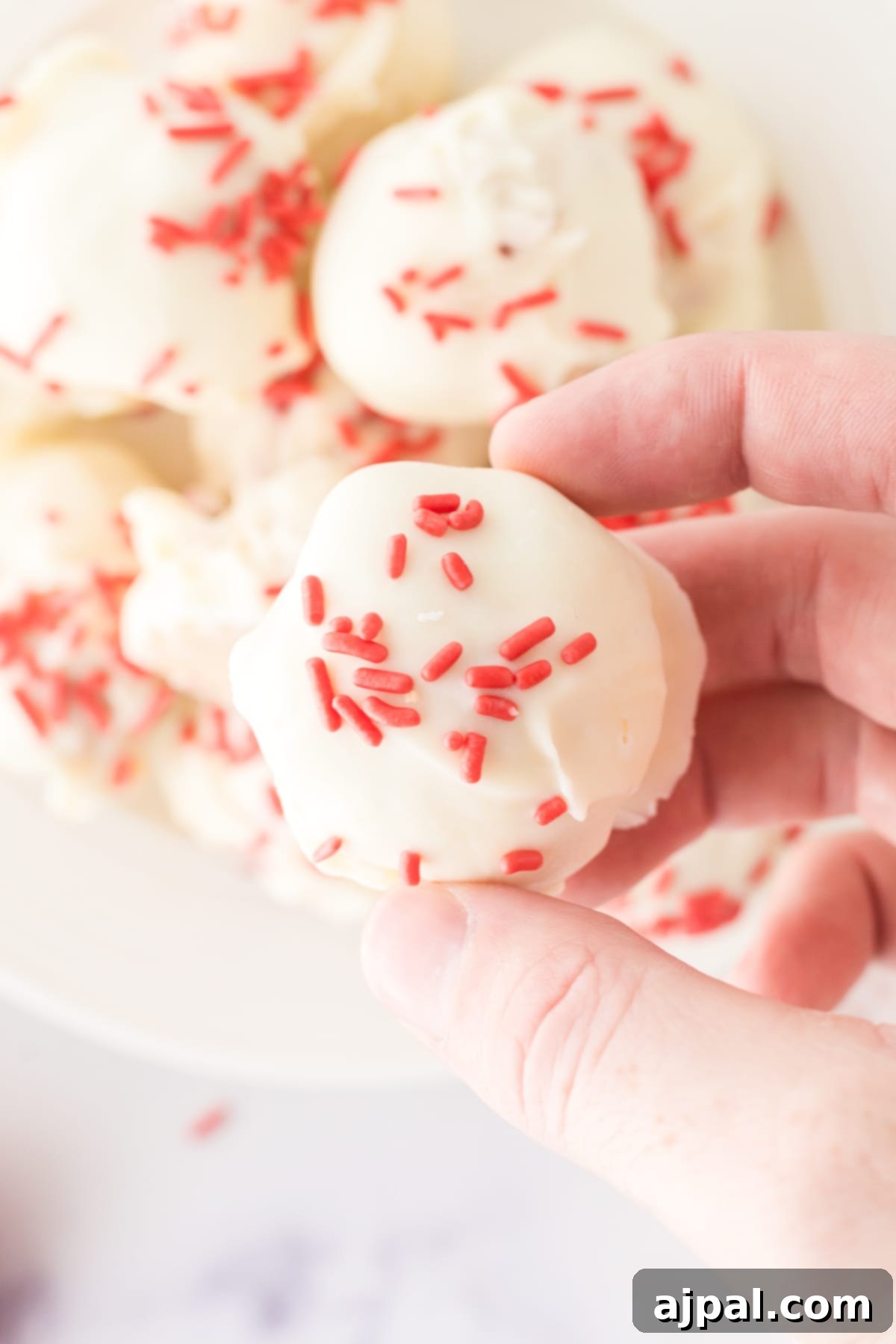 A hand holds a single white chocolate-dipped strawberry shortcake truffle, adorned with pink sprinkles. The truffle appears moist and inviting.