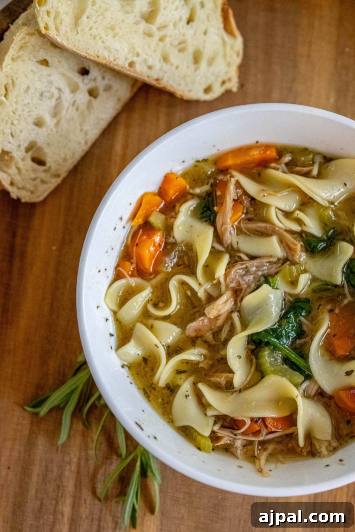 Hearty Slow Cooker Turkey Noodle Soup 11 A bowl of slow cooker turkey noodle soup, brimming with egg noodles, turkey, spinach, carrots, and celery, accompanied by crusty bread on a wooden surface.