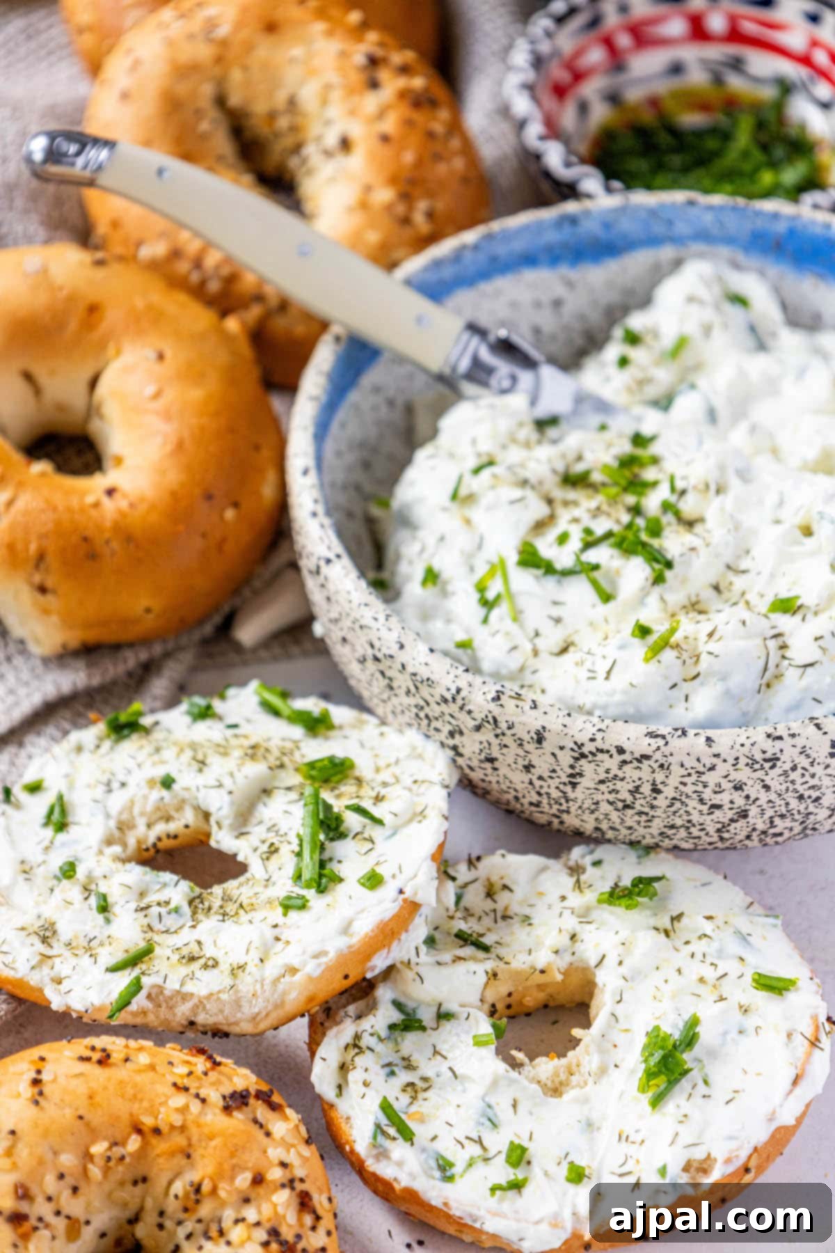 Several everything bagels arranged on a table, with one generously covered in creamy dill schmear and garnished with fresh chives and pickle seasoning.