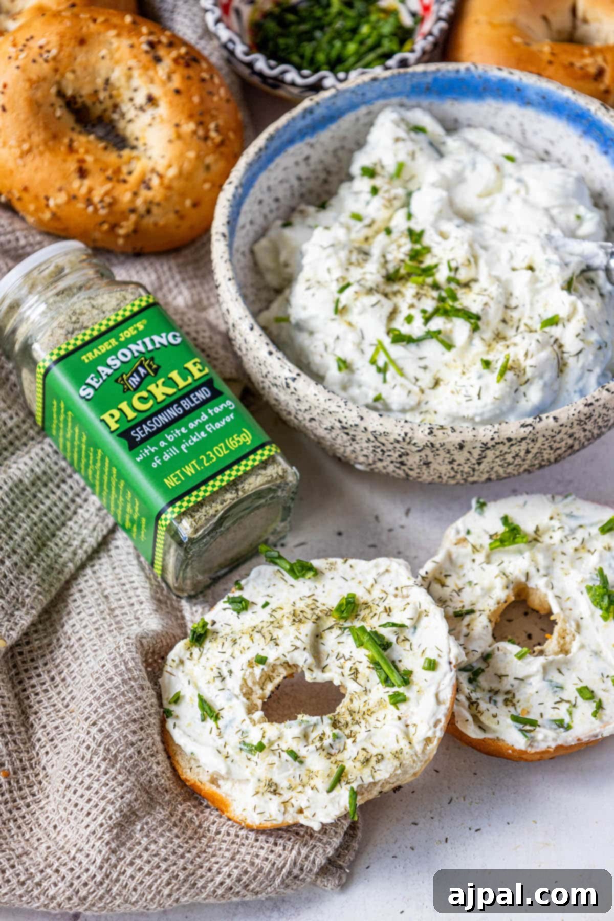 Two everything bagels generously topped with creamy garlic dill schmear, fresh chives, and pickle seasoning, sitting on a wooden surface.