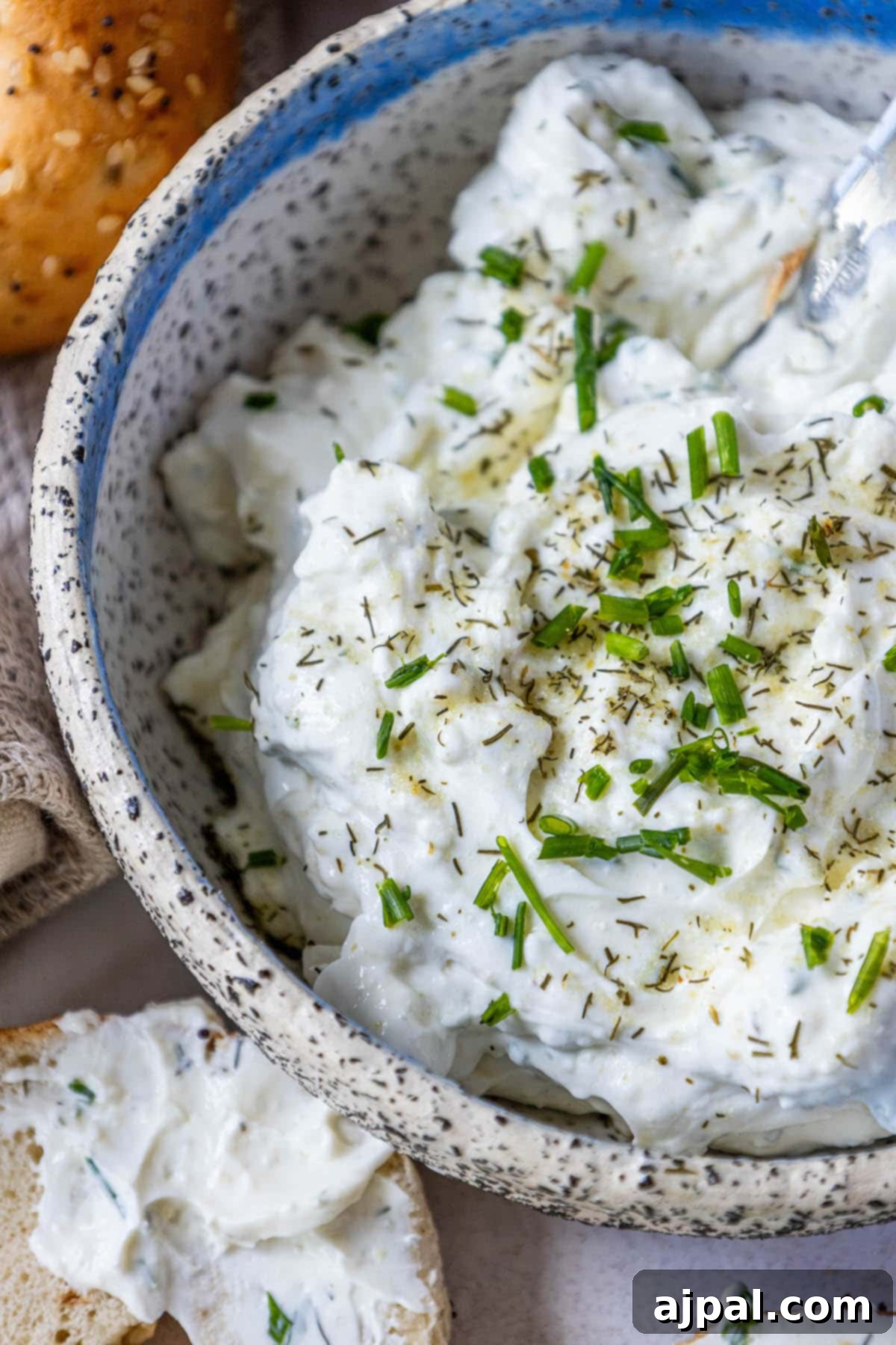 A bowl of creamy garlic dill schmear surrounded by various everything bagels, artfully arranged on a wooden table, inviting to be spread.