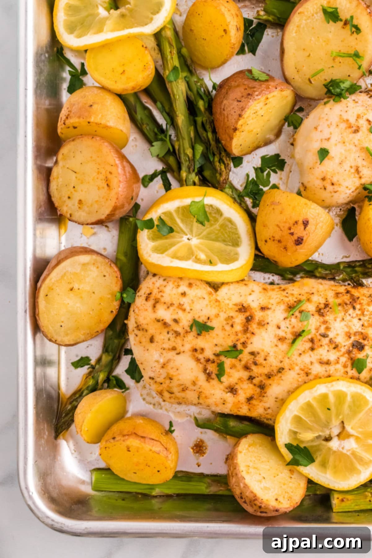 A sheet pan with perfectly baked lemon garlic chicken breasts, roasted baby potatoes, vibrant green asparagus, and fresh lemon slices.