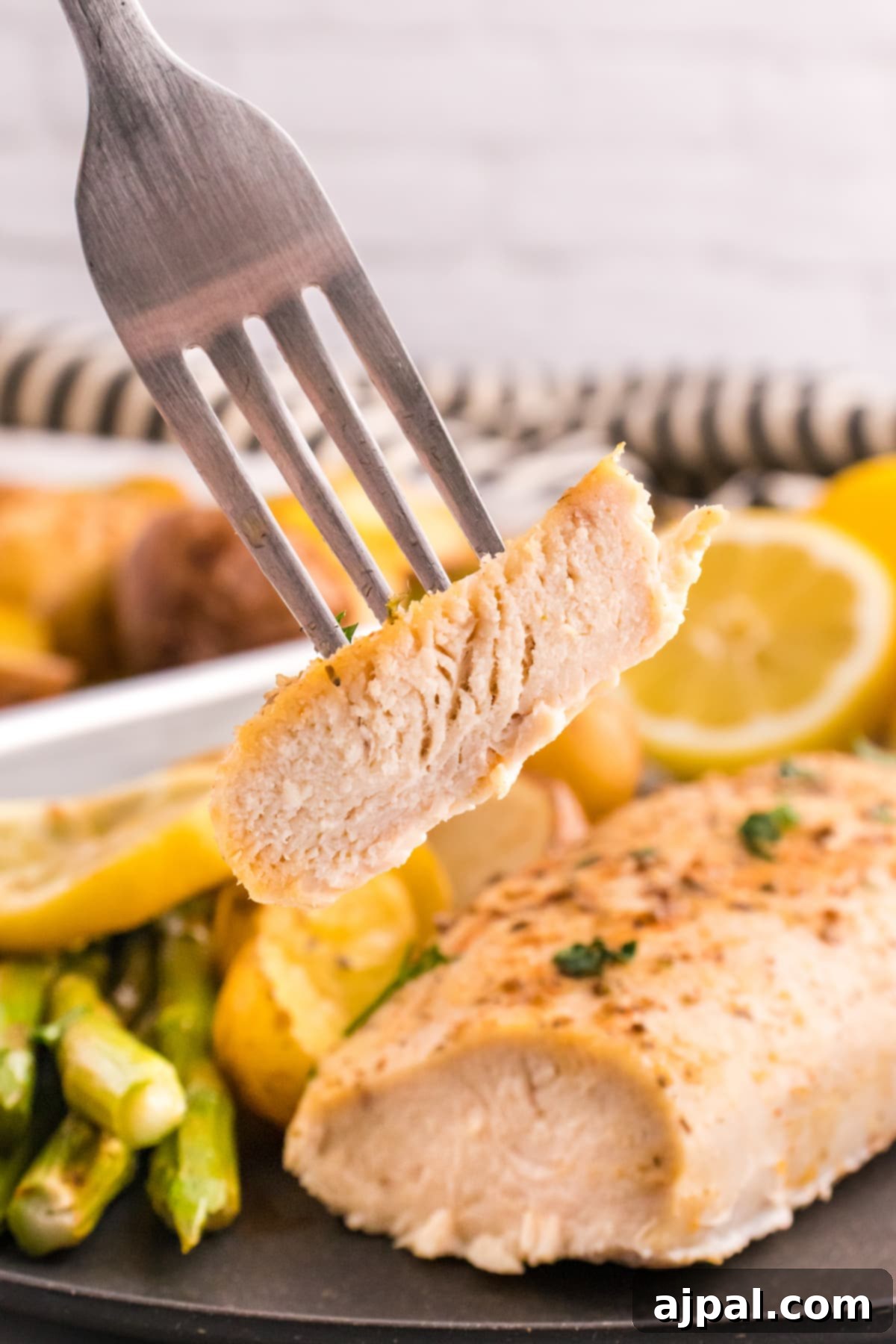 A fork holding a piece of lemon garlic chicken over a plate with baked chicken breasts, potatoes, lemon slices, and asparagus, ready to be enjoyed.