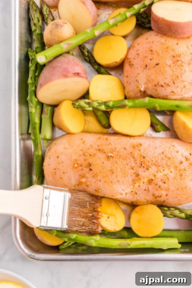A cooking brush applying a generous coat of lemon garlic marinade onto raw chicken and vegetables spread on a baking sheet.