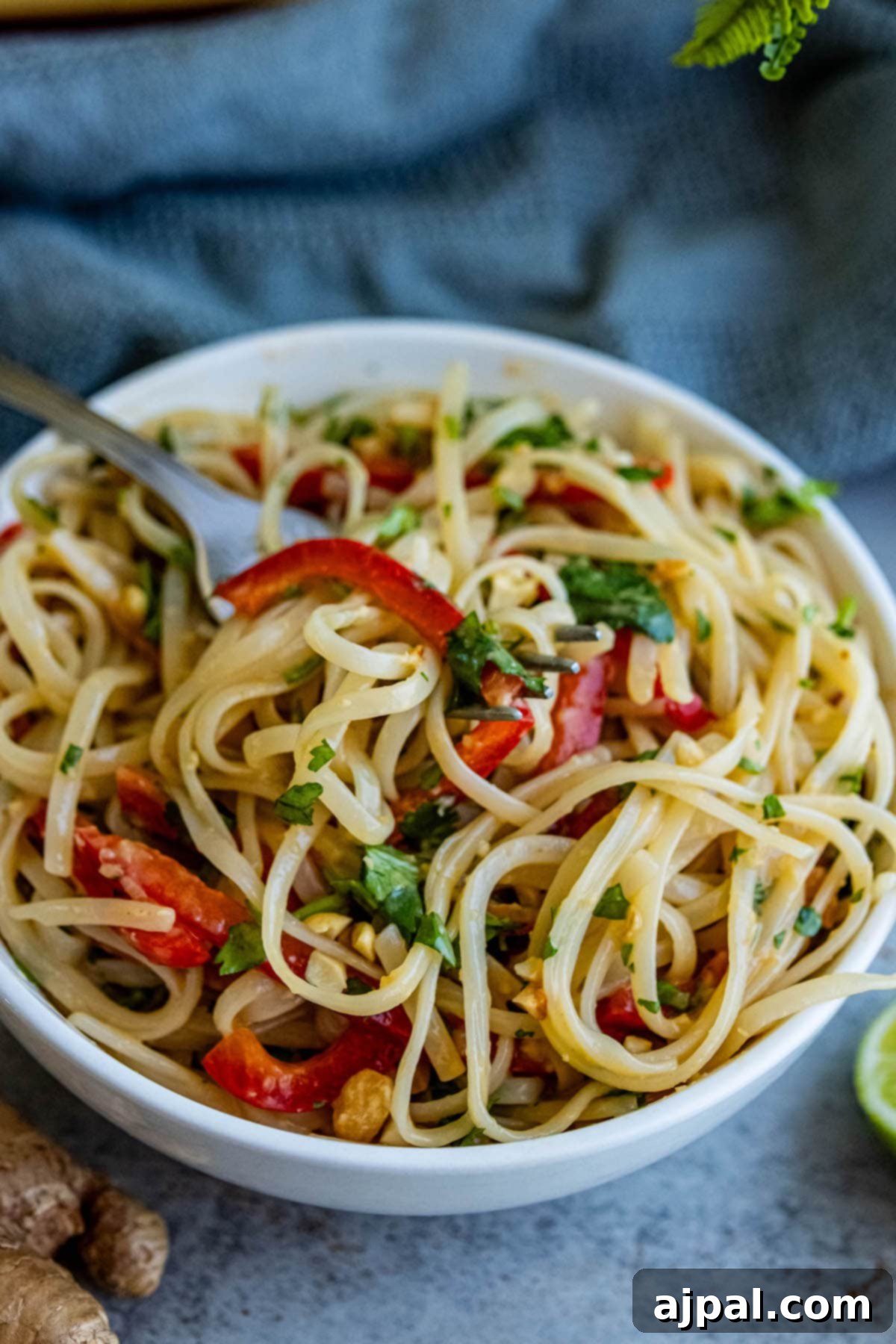Noodles with dressing, peppers, peanuts, and cilantro, limes around it, presented in an elegant white bowl.