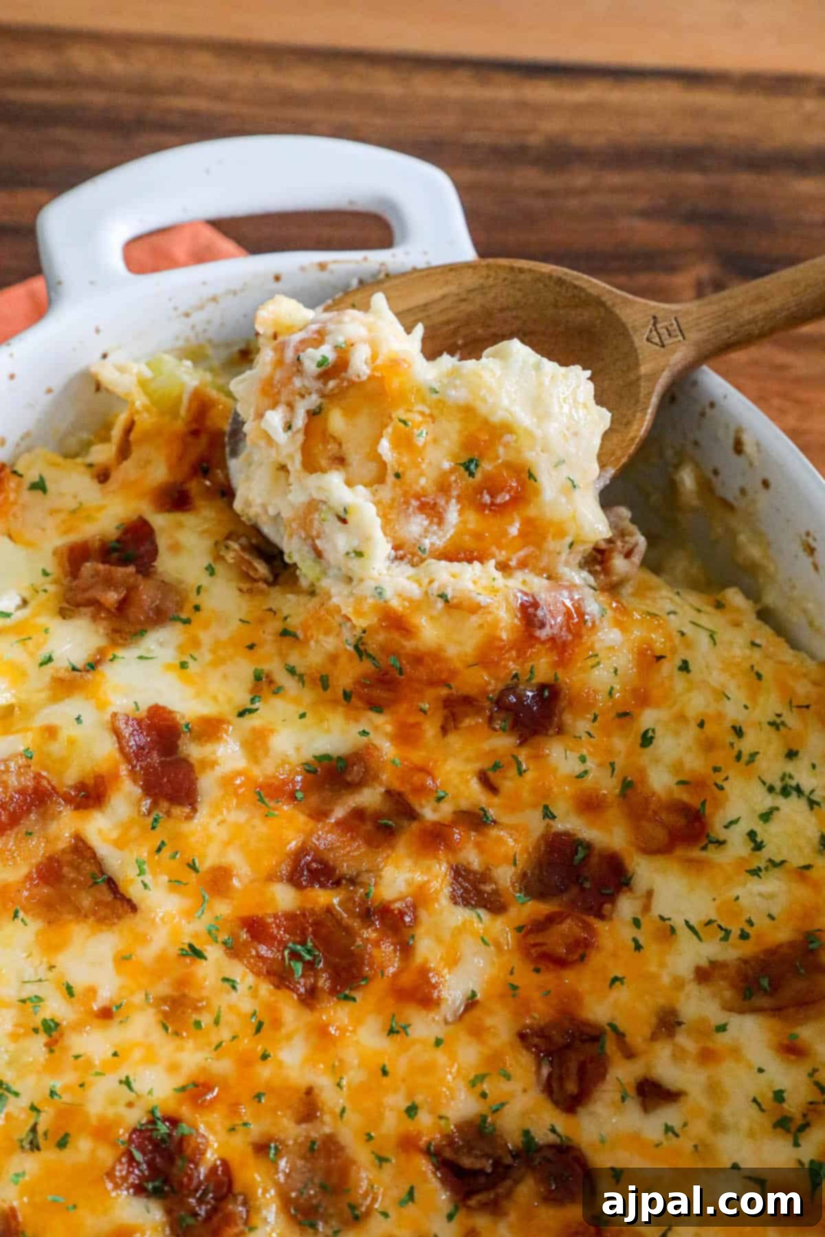 A serving of cheesy mashed potatoes, topped with crispy bacon, in a casserole dish alongside a wooden spoon.
