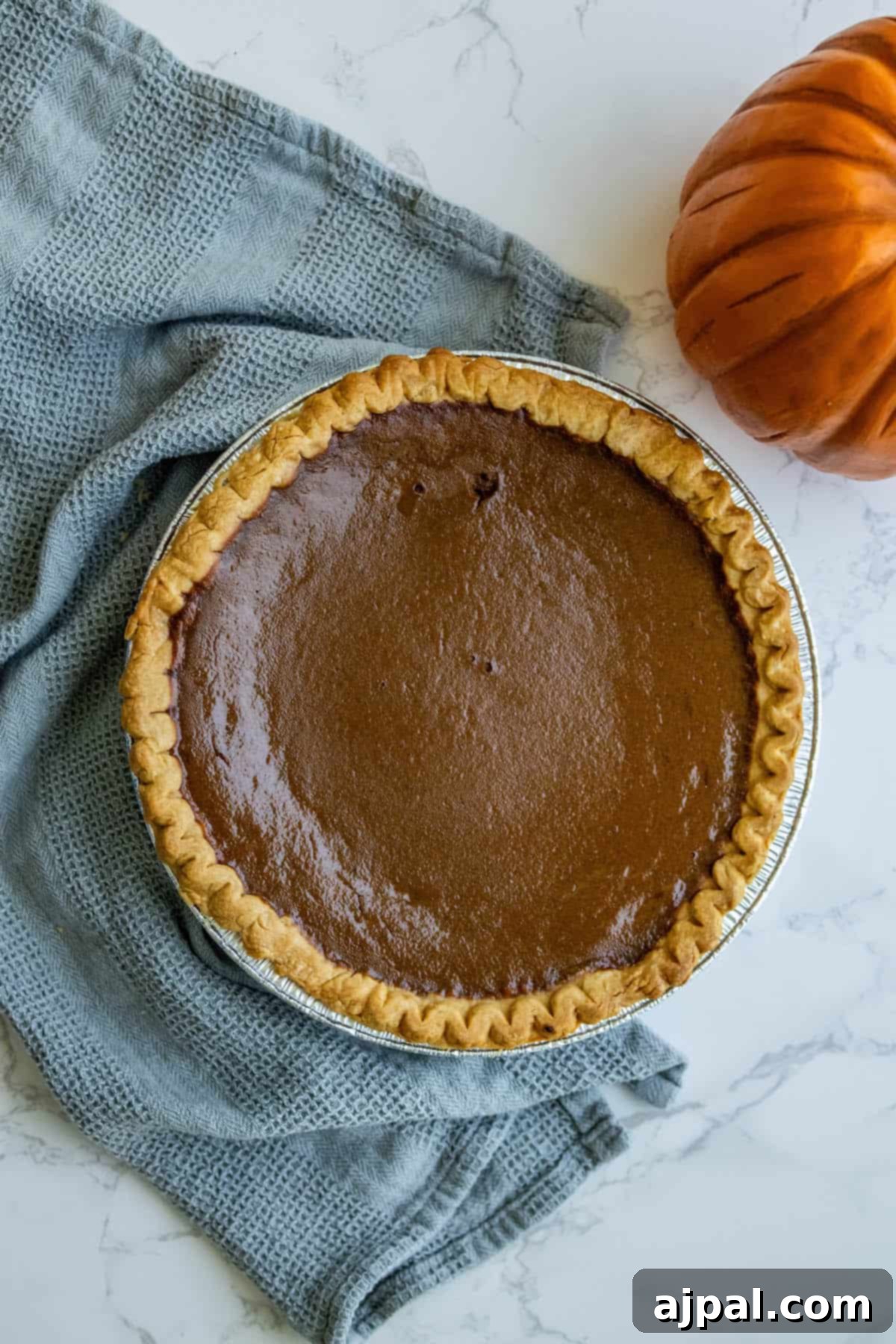 A golden-brown pumpkin pie sitting on a wooden table, next to a small orange pumpkin, evoking a warm, autumnal feel.