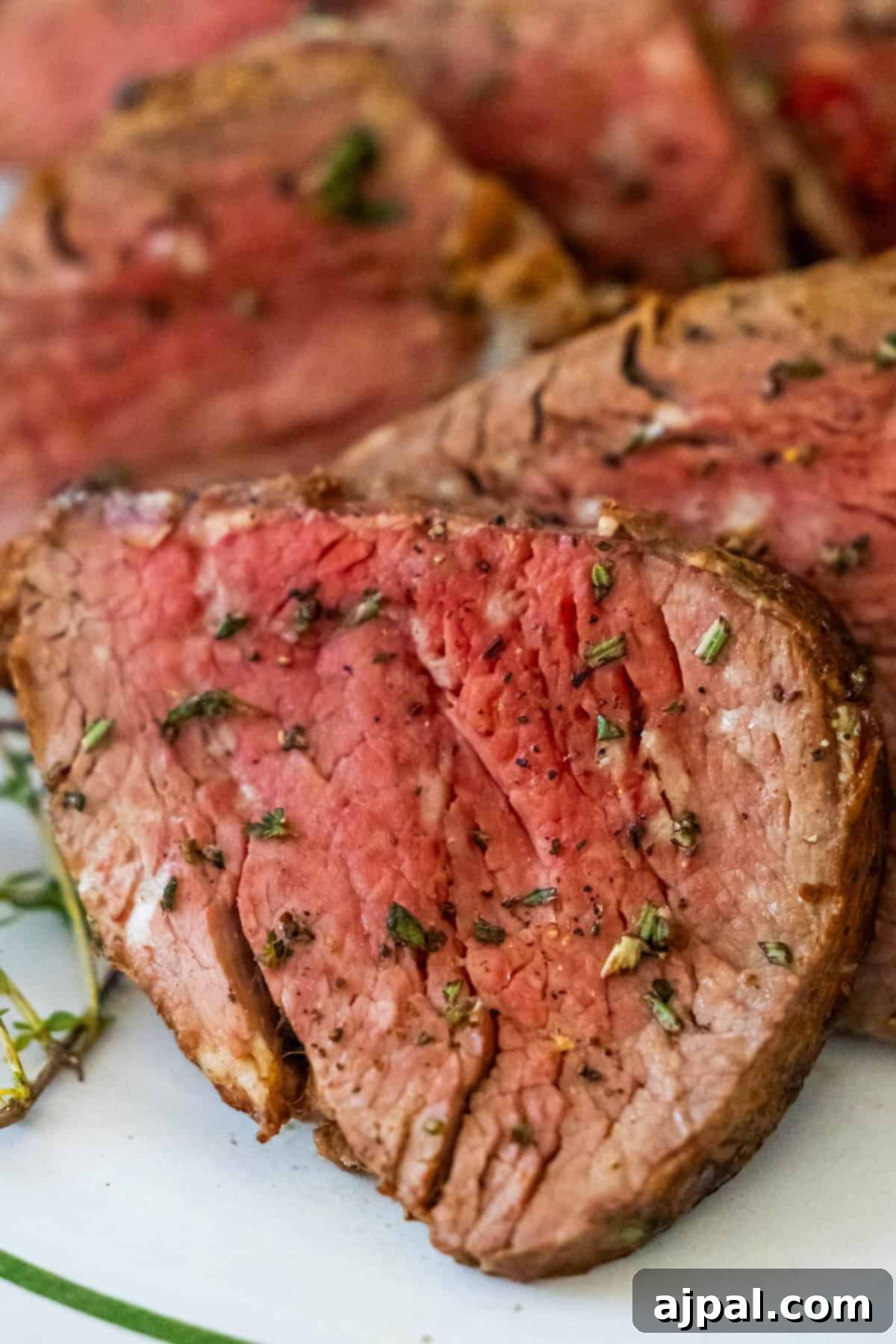 A close-up of sliced medium-rare beef tenderloin, seasoned with herbs, elegantly arranged on a white plate.